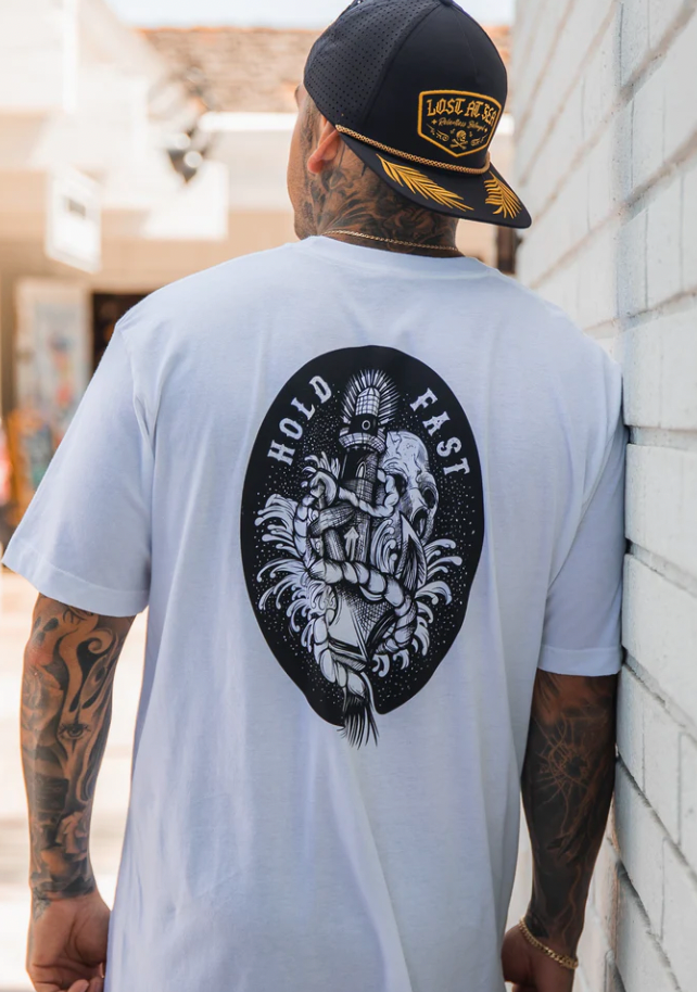 A tattooed individual wears a Relentless Betrayals Hold Fast White T-Shirt, featuring a hand clutching a lighthouse in black and white with HOLD FAST text. They pair it with a cap and stand against a white brick wall.