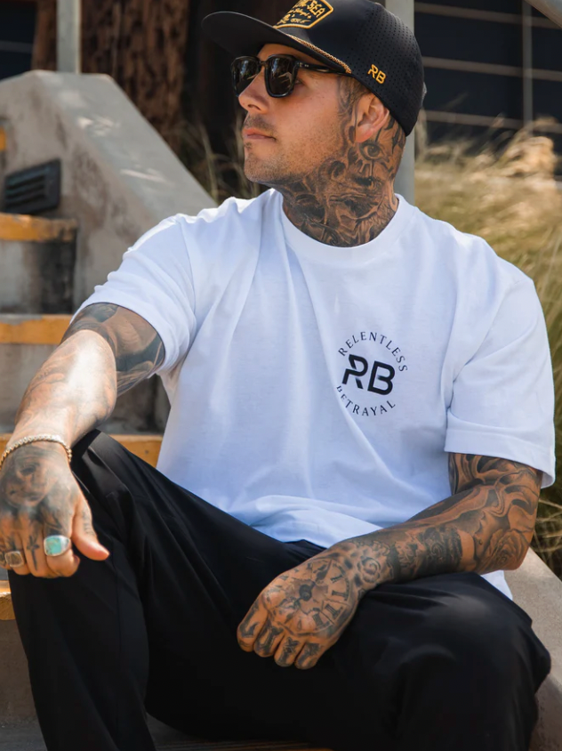 A tattooed individual sits on outdoor steps, wearing a Relentless Betrayal Hold Fast White T-Shirt in fine jersey cotton. Complementing the look, they sport a black cap, sunglasses, black pants, and a ring, casting a neutral gaze aside for an intriguing casual yet unisex style.