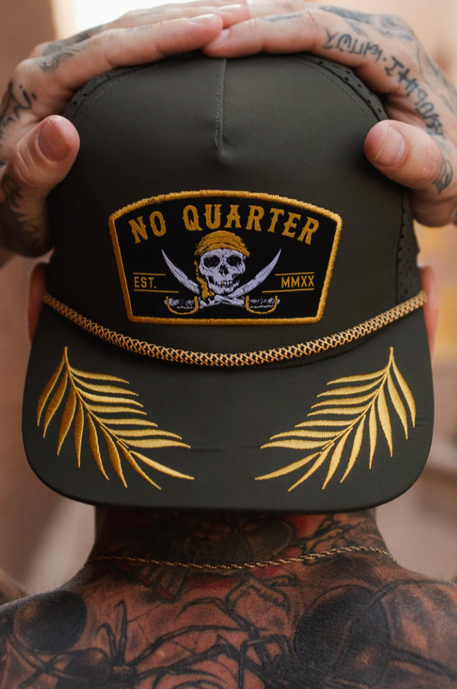 A person with tattooed hands and neck holds their head, wearing the Relentless Betrayal No Quarter Olive Snapback, featuring a skull and crossed swords emblem, yellow palm leaves embroidery, a chain decoration, and No Quarter Est. MMXX, all in a water-resistant design.