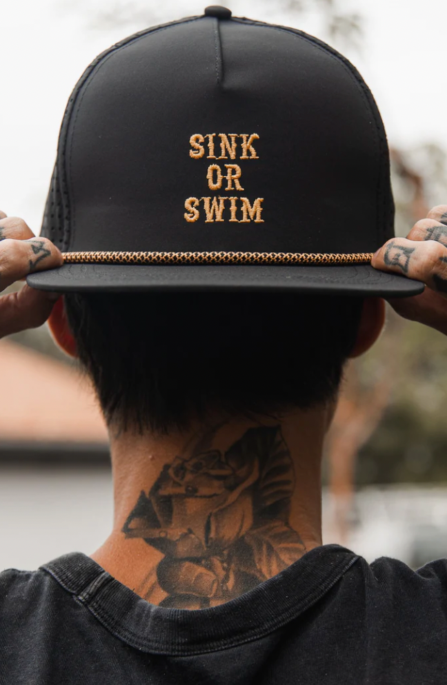A person adjusts their Relentless Betrayal Sink or Swim Blackout Snapback featuring gold chain detail and SINK OR SWIM in gold lettering. Tattooed hands and visible neck tattoos add an edgy vibe to the water-resistant hat against a blurred background.
