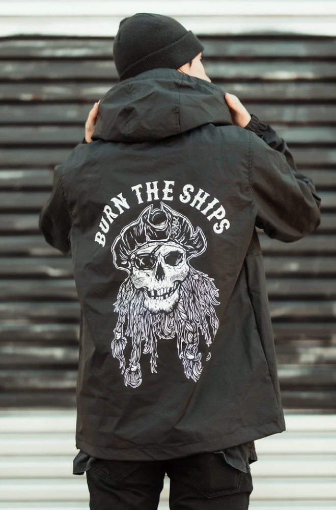 An individual adjusts the hood of their Burn the Ships Pullover Rain Jacket by Relentless Betrayal. The black unisex jacket features a skull in a pirate hat with braids graphic and BURN THE SHIPS above it. Its water-resistant coating stands out against the striped backdrop behind them.
