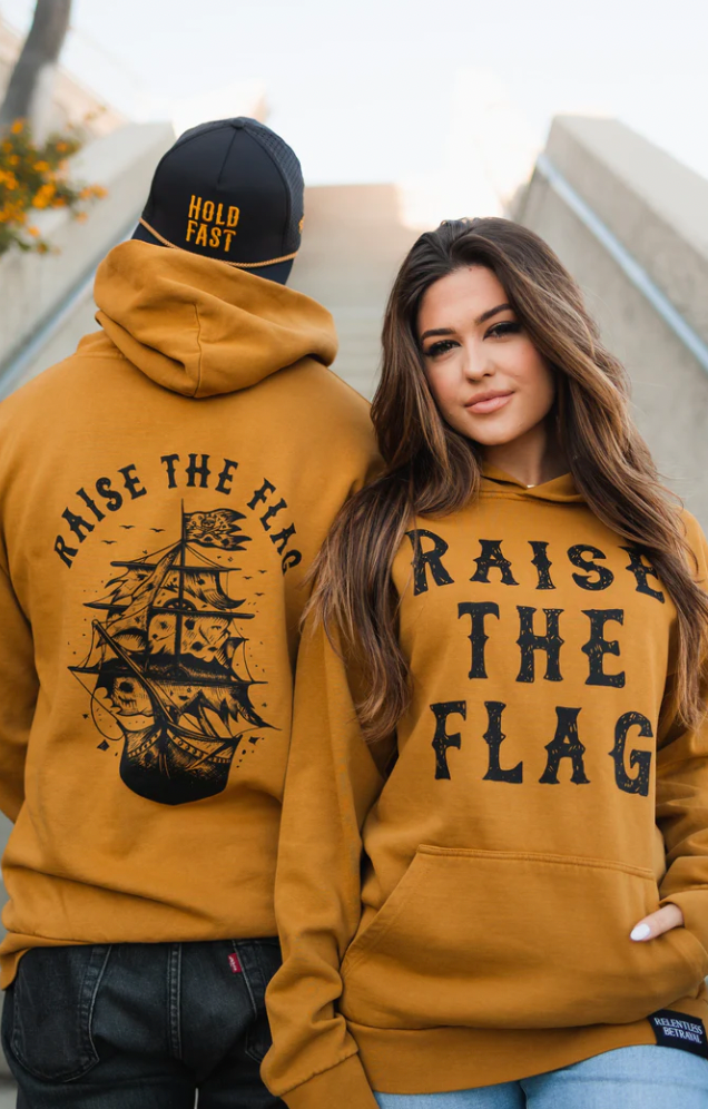 A man and woman sport matching Relentless Betrayal Raise the Flag premium toast unisex hoodies, each with RAISE THE FLAG on them. The mans back showcases a pirate ship graphic, and he wears a black cap stating HOLD FAST. They stand on steps outdoors, facing different directions.