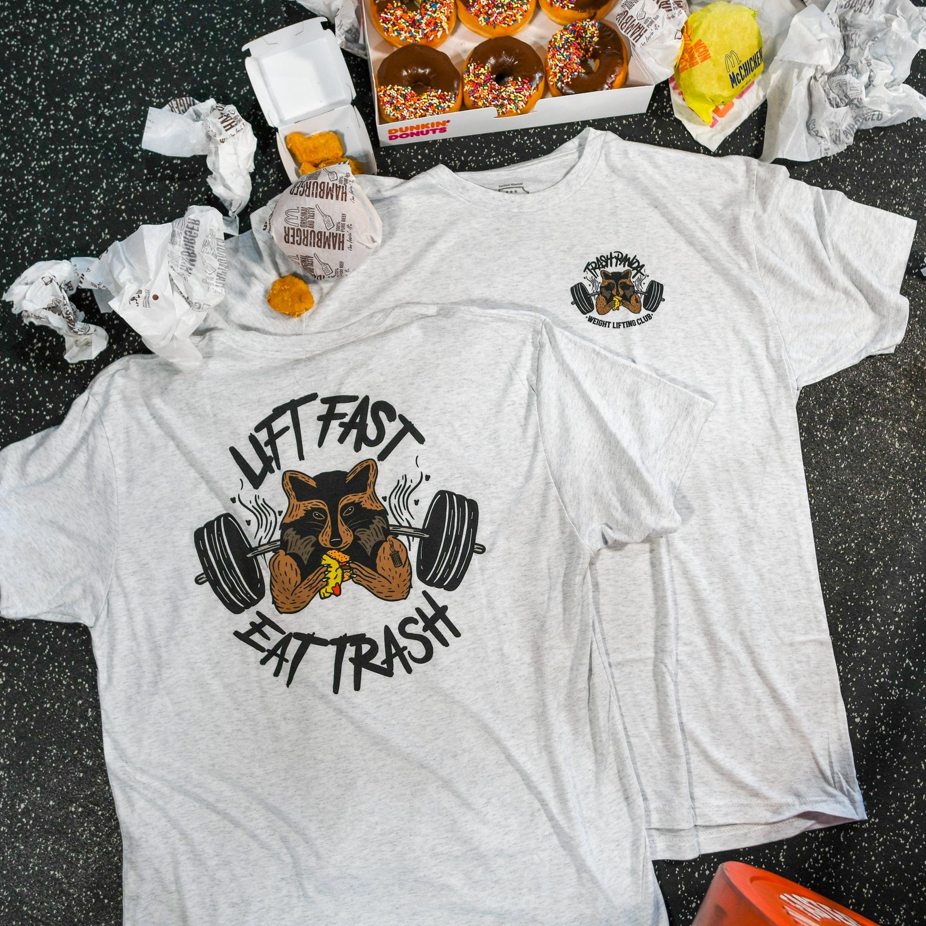 The Trash Panda Weight Lifting Club Tee by Tango Charlie Apparel features a gray tri-blend design with a trash panda lifting weights and eating a burger. Surrounding the Lift Fast, Eat Trash text are fast food items like donuts, wrappers, and nuggets on a speckled surface.