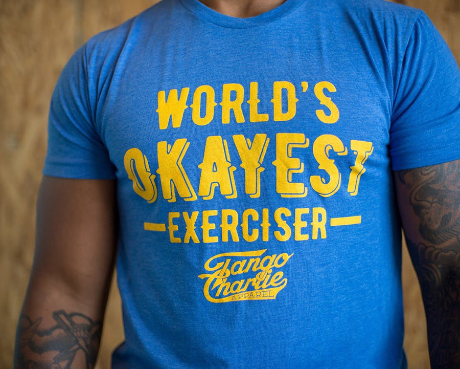 A person wears a comfy blue cotton/poly blend tee with yellow WORLDS OKAYEST EXERCISER text from Tango Charlie Apparel, set against a blurred wooden background with tattoos on their arm.