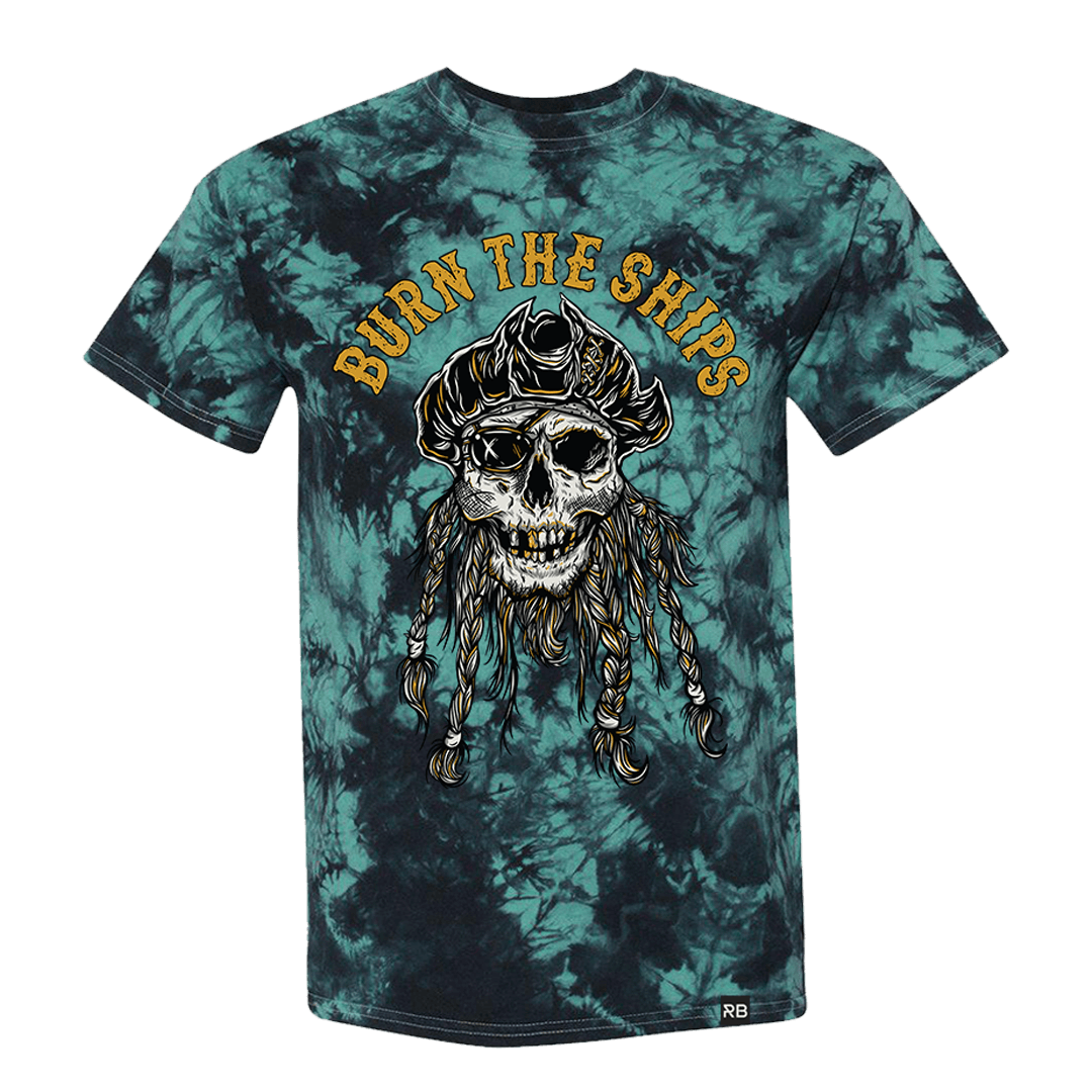 The Burn The Ships Tie Dye Tee by Relentless Betrayal features a pirate skull with a tattered hat and braids. Burn the Ships appears in orange above the skull, set against a blue and black tie-dye design. Made from fine jersey cotton, it offers an edgy style with ultimate comfort.
