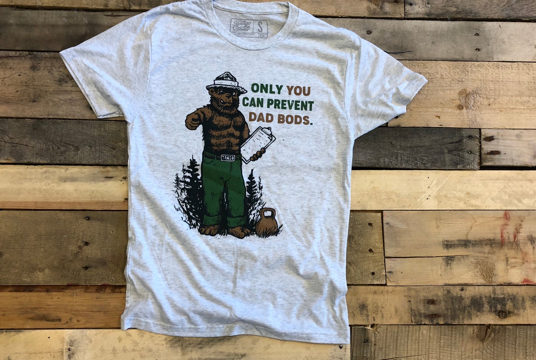The Only You Can Prevent Dad Bod T-Shirt by Tango Charlie Apparel features a bear in green pants with a clipboard and the humorous slogan ONLY YOU CAN PREVENT DAD BODS, set against forest trees on a gray tri-blend fabric atop a wooden background.