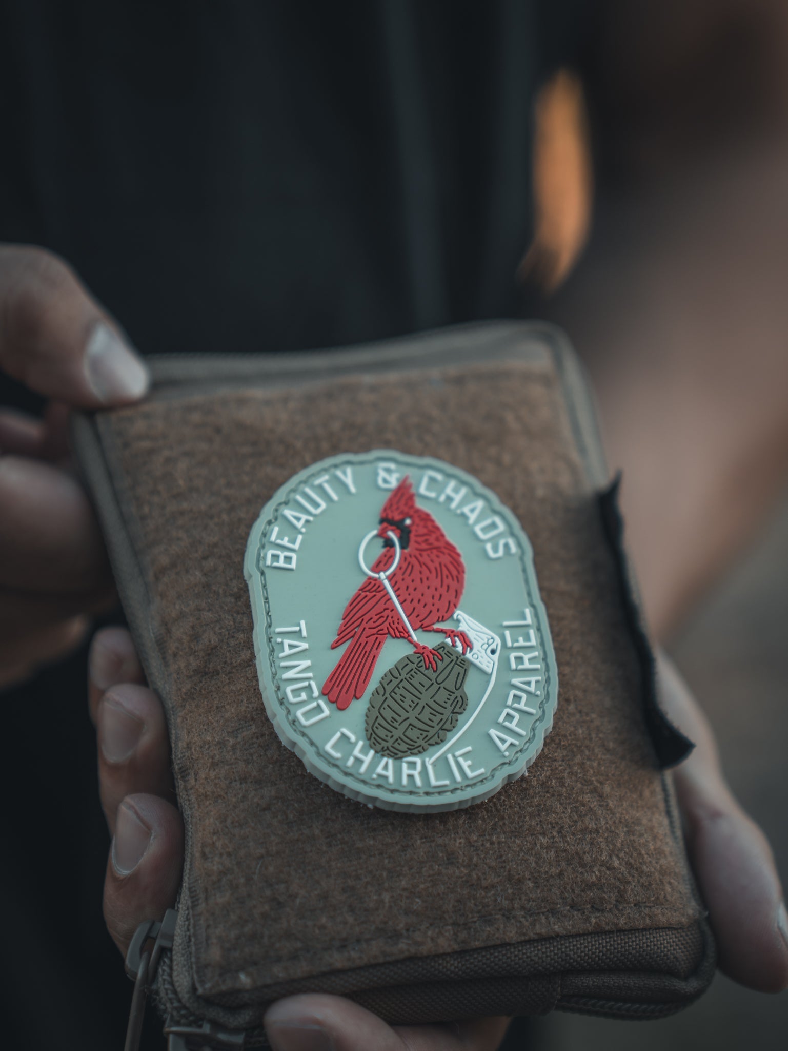 A person holds a brown pouch with a patch that reads BEAUTY & CHAOS TANGO CHARLIE APPAREL and features a red bird perched on a green dog tag.