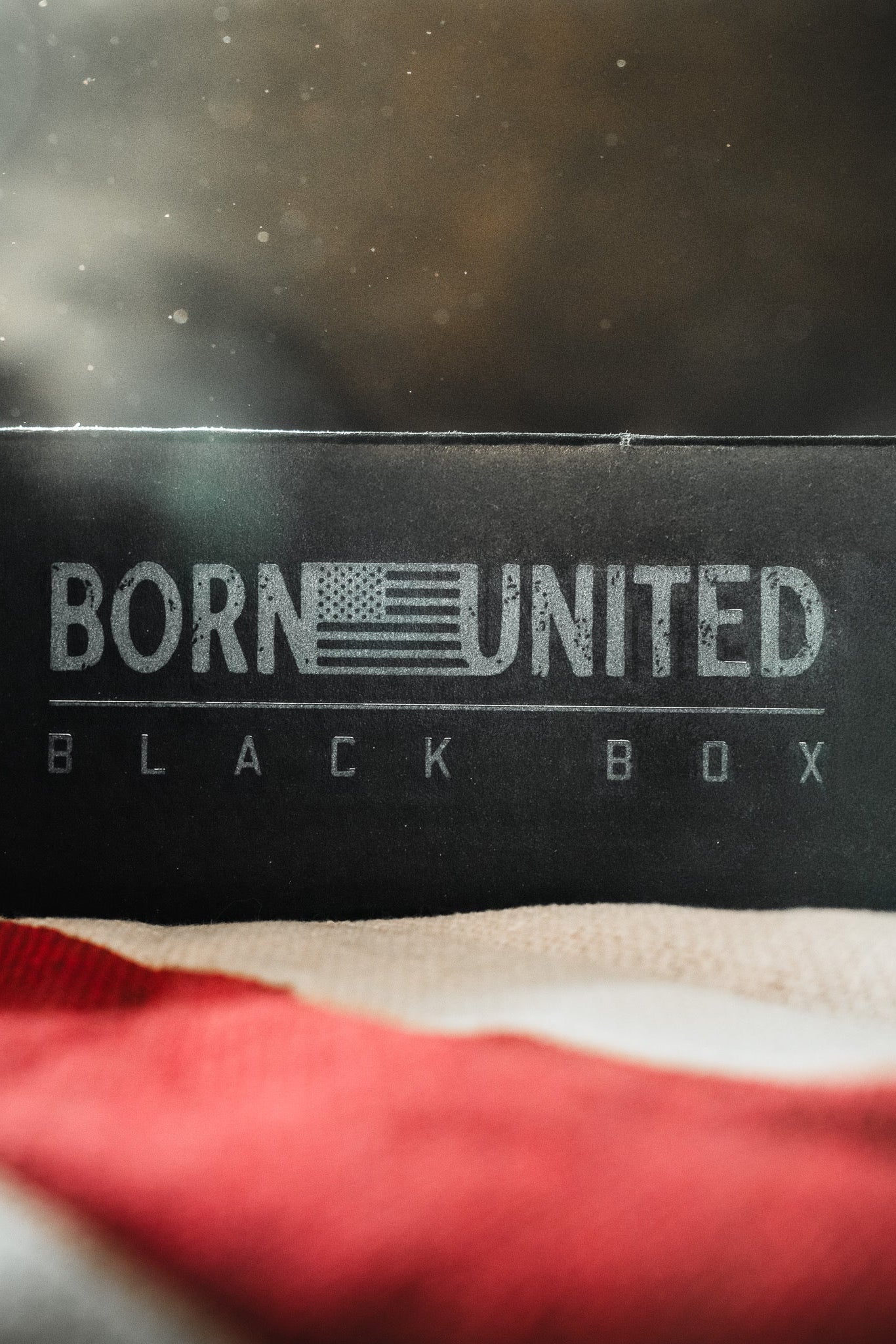A black box with the text BORN UNITED featuring a stylized American flag design, and BLACK BOX written below. An out-of-focus American flag is in the foreground.