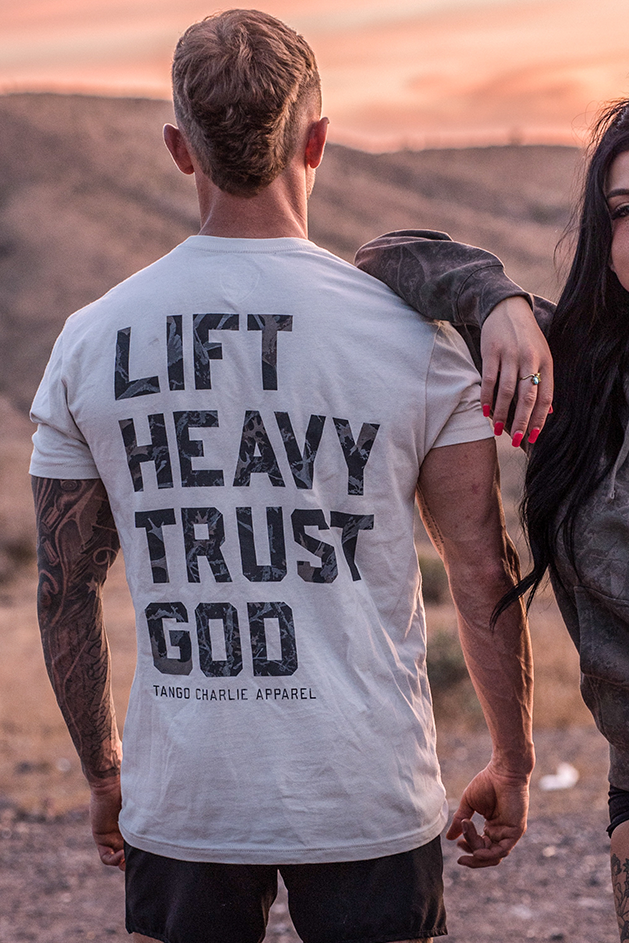 A muscular person standing outdoors with their back to the camera, wearing a shirt that says LIFT HEAVY TRUST GOD. Another person stands beside them with an arm draped over their shoulder.