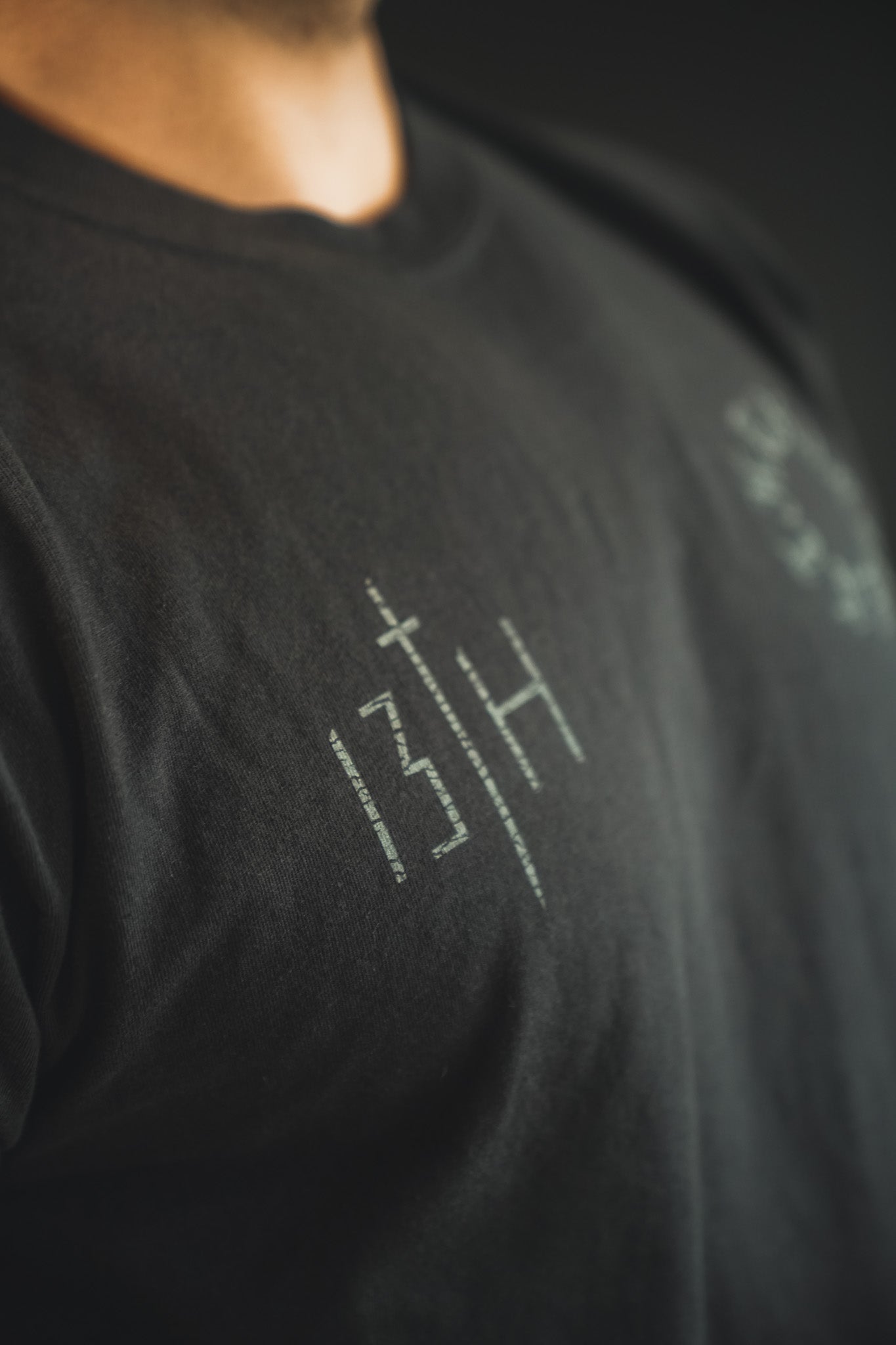 Close-up of a person wearing a black T-shirt with minimalist white text and symbols, including the numbers 13 and H separated by a vertical line. The background is dark and out of focus.