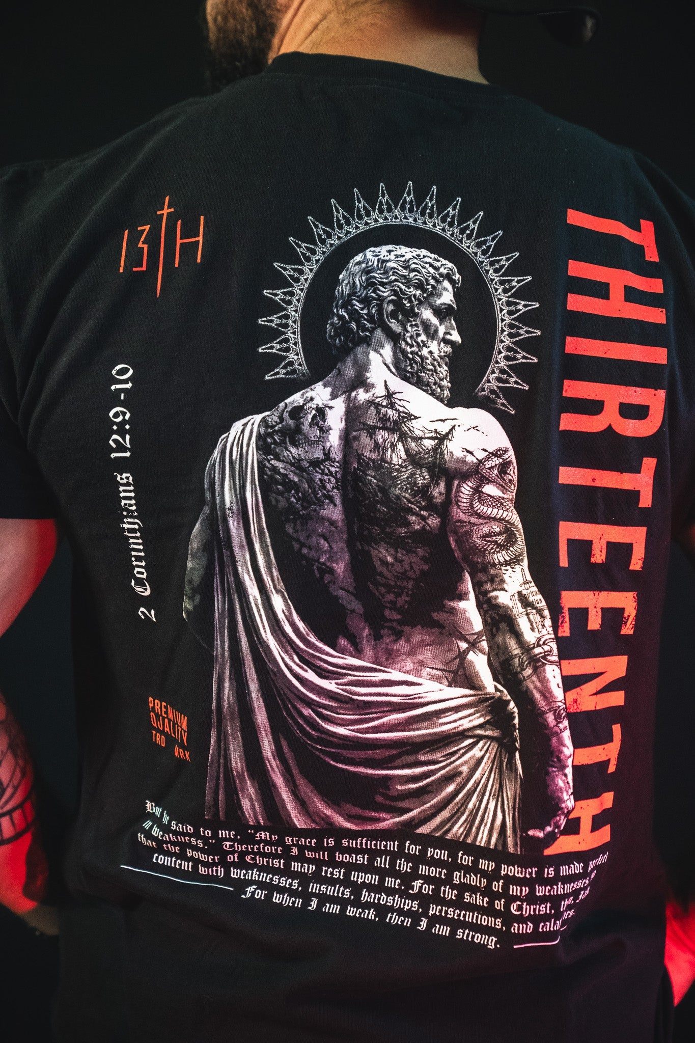 A person wearing a black T-shirt with a large graphic of a Roman figure, a halo, text THIRTEENTH,” 2 Corinthians 12:9-10, and red and white lettering on the back. Only their back is visible.