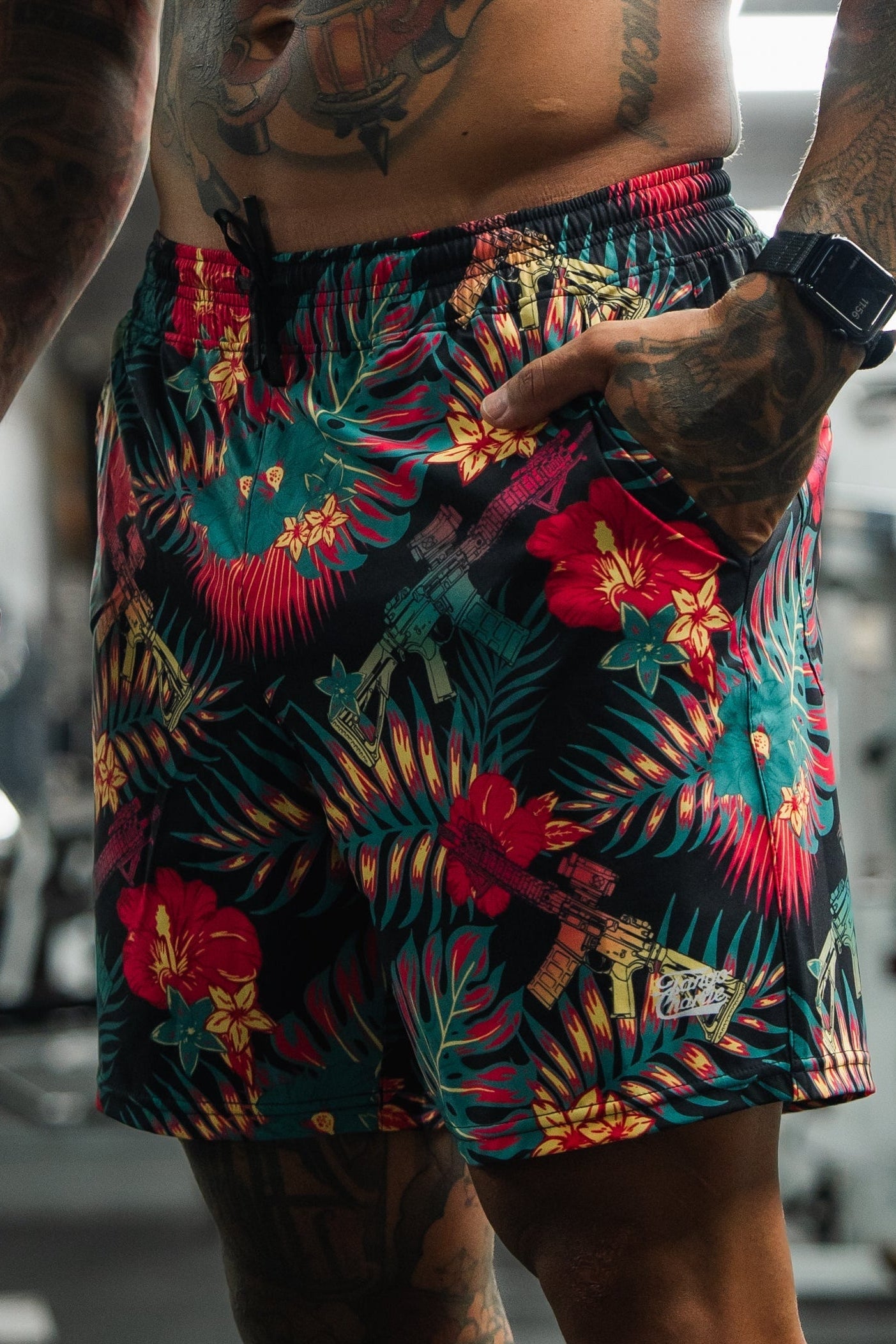 A person wearing colorful tropical-print shorts with red, yellow, and green leaves and flowers, also featuring illustrated rifles in the design. The person has tattooed arms and is standing indoors.