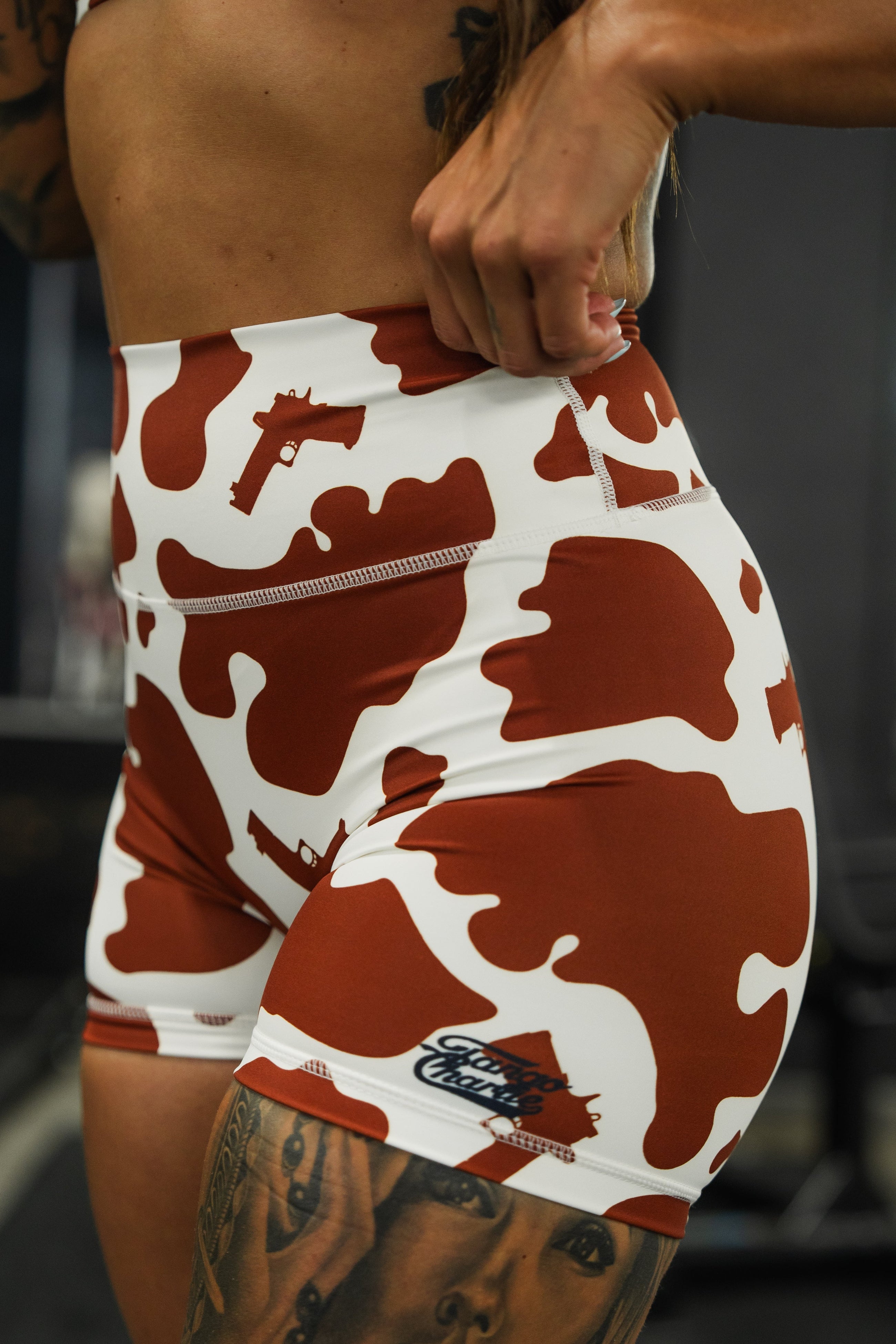 A person wearing red and white camouflage shorts with gun shapes and a Gritty Threads logo, showing their lower torso and hand adjusting the waistband.