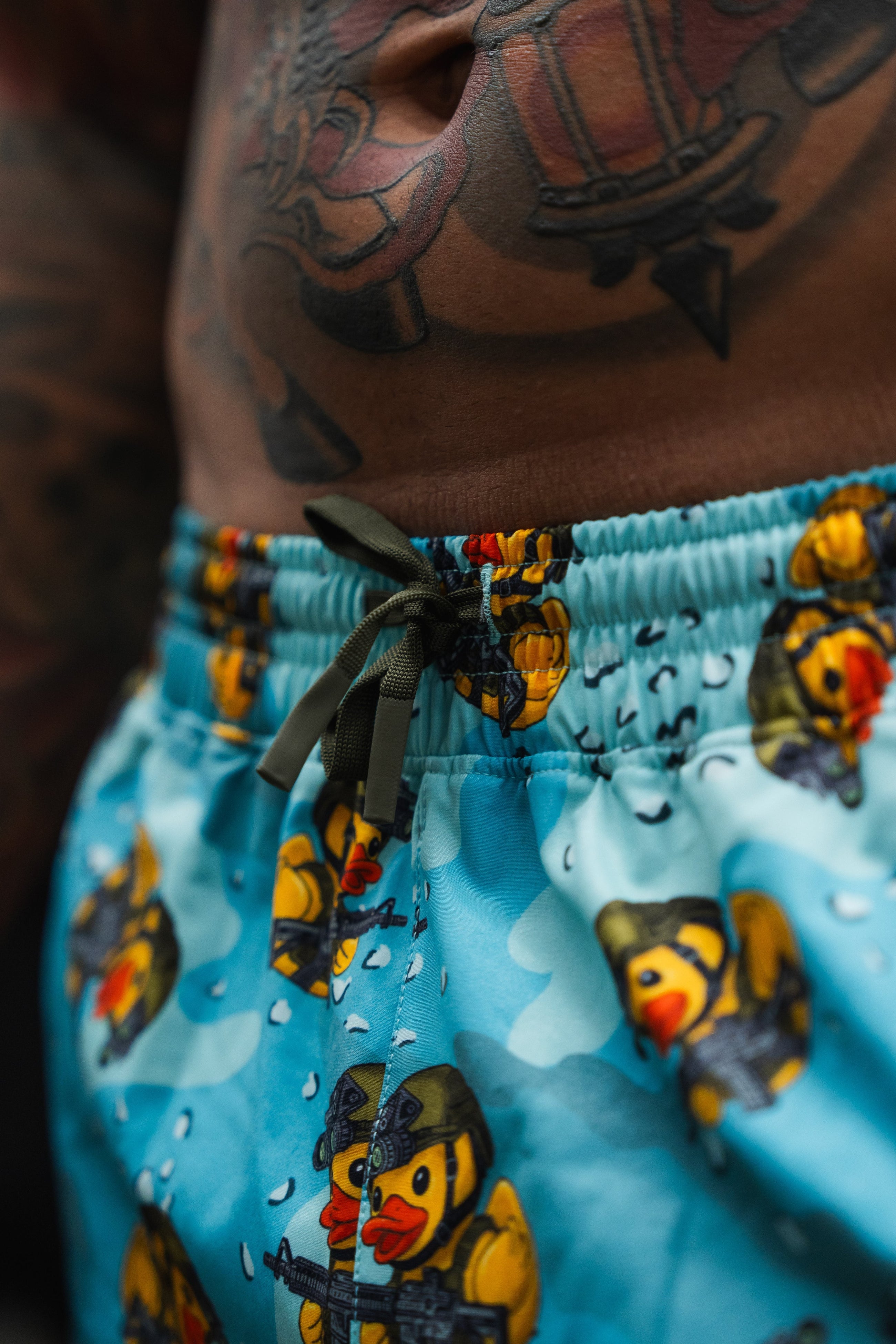 Close-up of a persons torso showing detailed tattoos and blue shorts with cartoon rubber ducks dressed in military gear, holding guns, and wearing helmets. The shorts have a black drawstring.