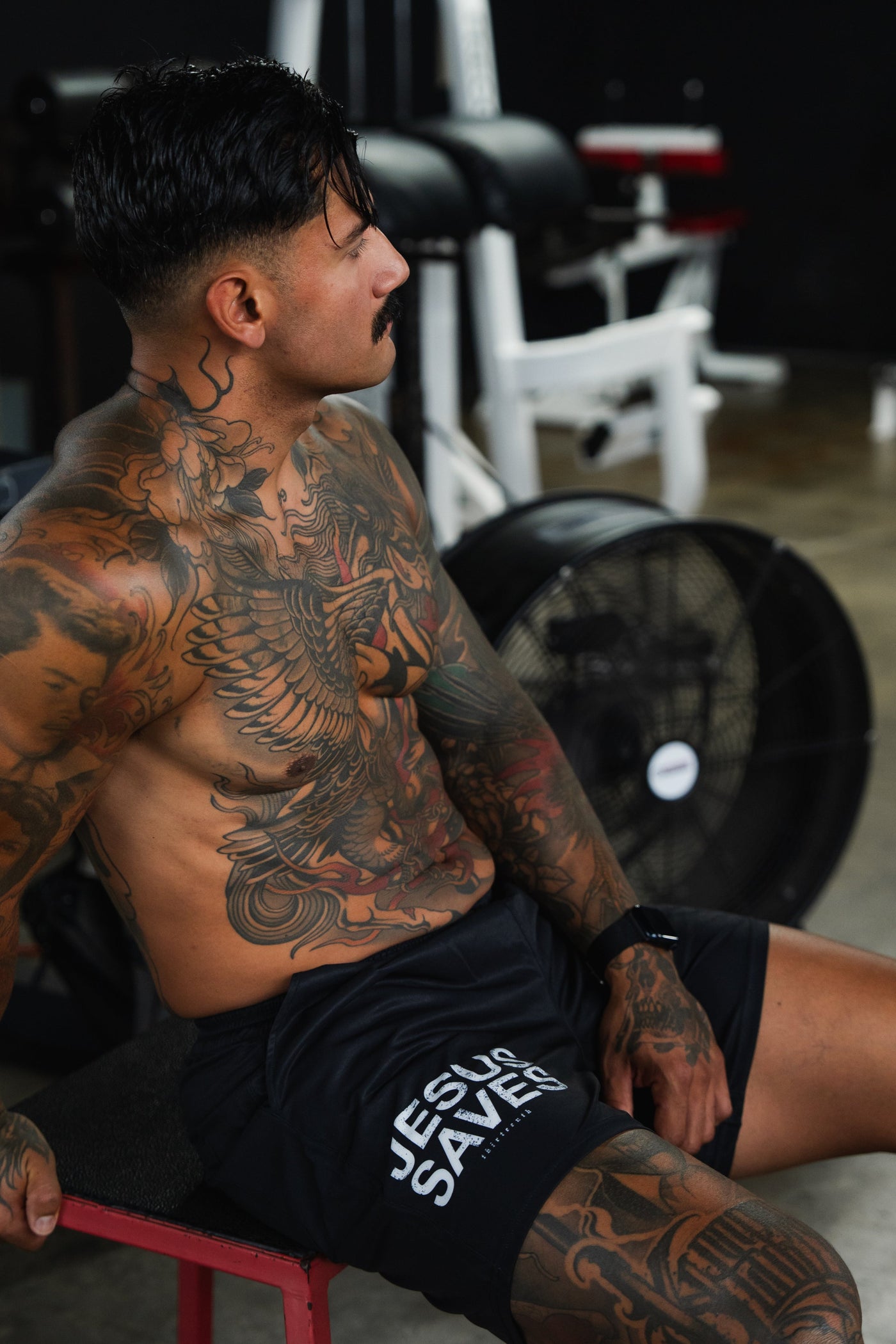 A muscular, tattooed man with dark hair sits on a bench in a gym, wearing black shorts that say JESUS SAVES. Gym equipment and a large fan are visible in the background.