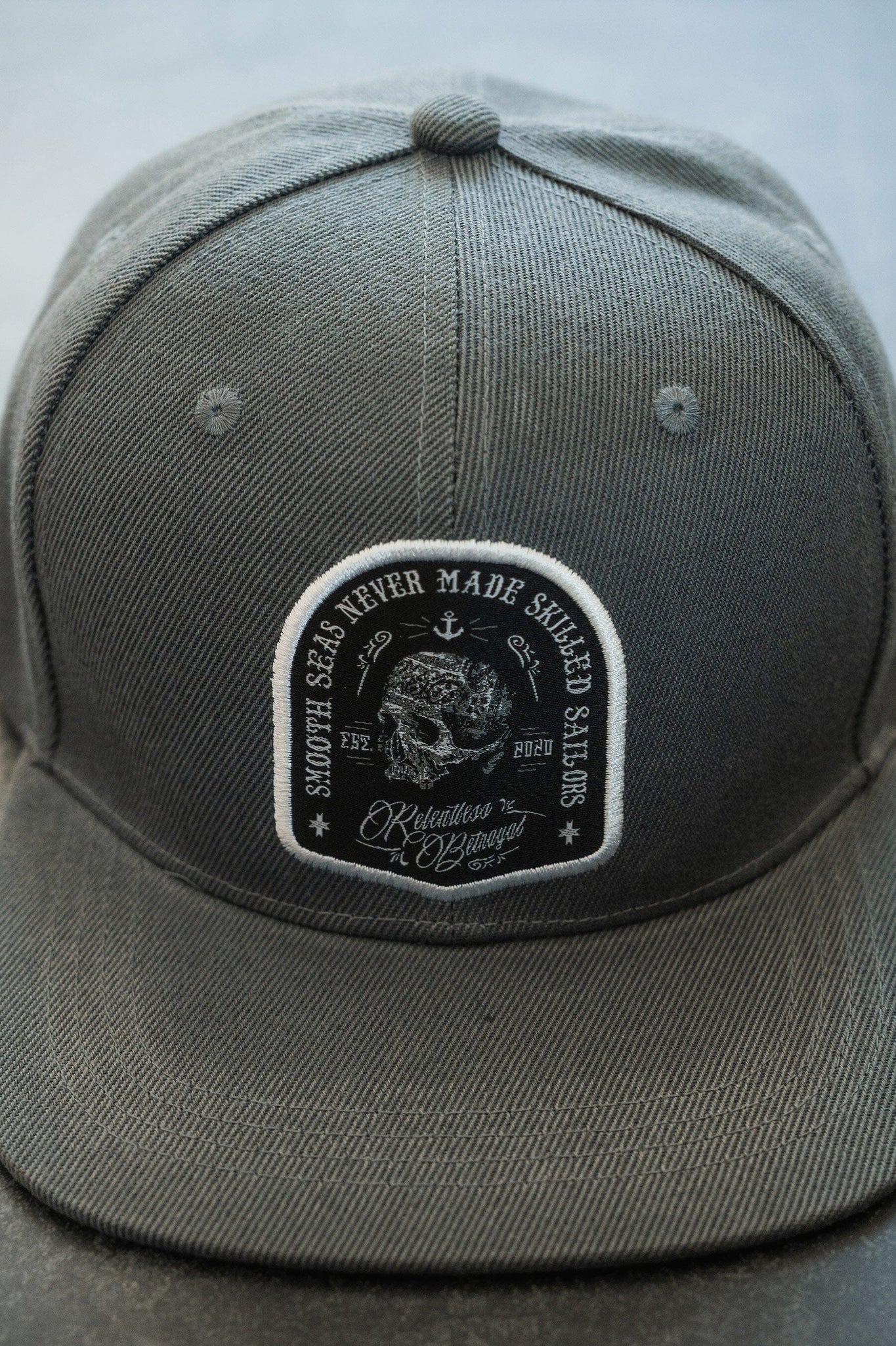 A gray baseball cap with a black and white patch featuring a skull, anchor, and text that reads Smooth seas never made skilled sailors and Relentless & Unstoppable.