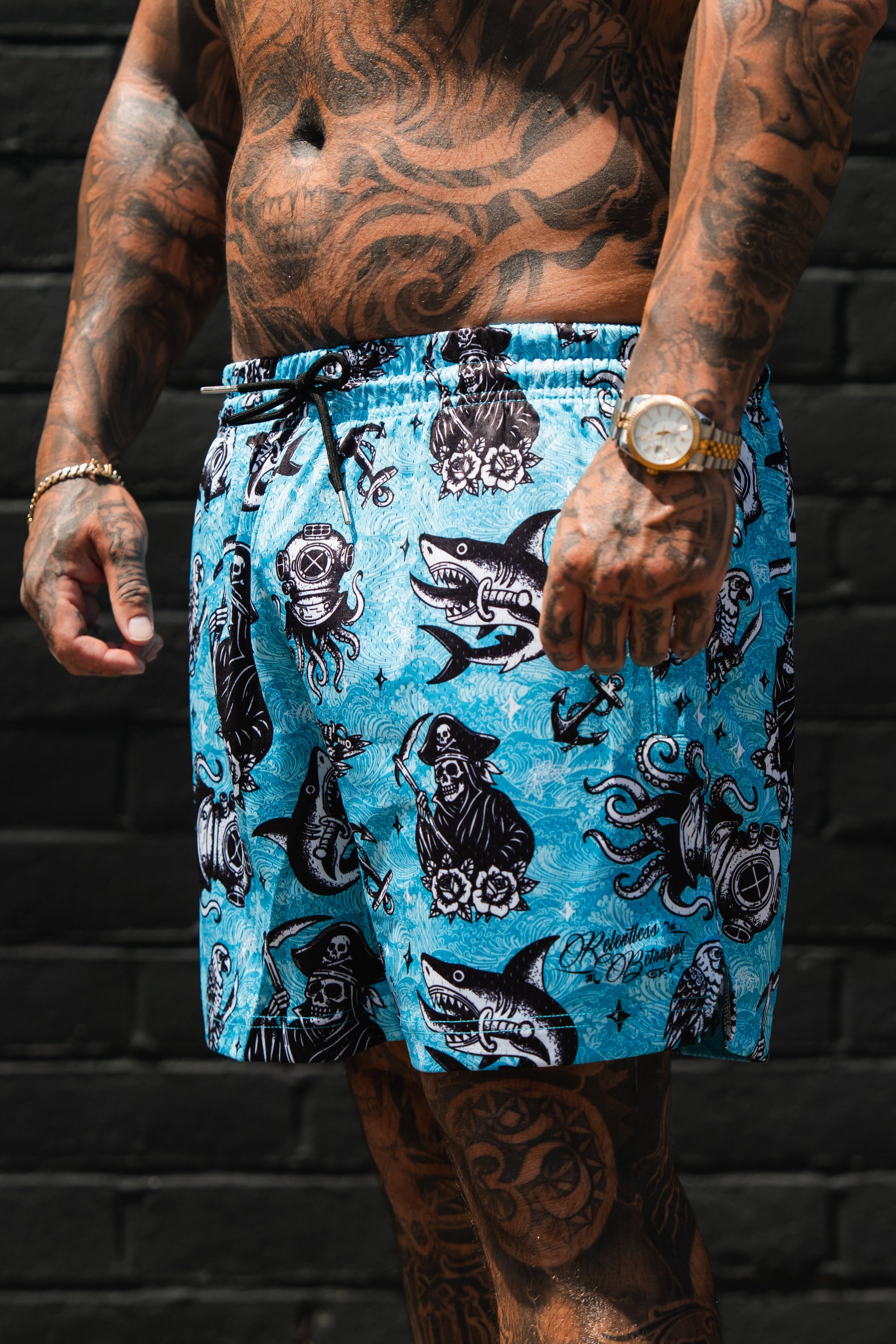 A tattooed man stands before a dark brick wall wearing Relentless Betrayal’s Premium Mesh Shorts | 5.5"- Davy Jones, featuring bold black and white nautical designs—sharks, mermaids, octopuses, and ships—for standout summer style.