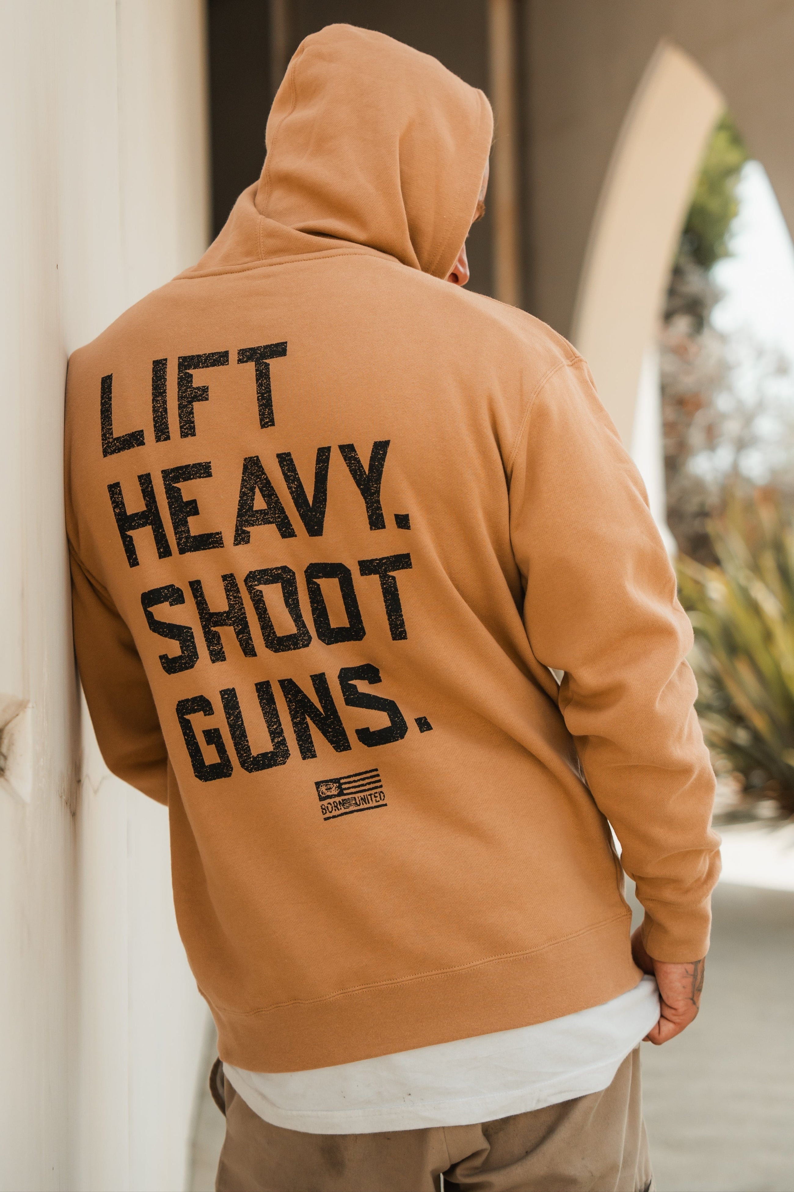 A person wearing a tan hoodie with the text LIFT HEAVY. SHOOT GUNS. printed in black letters on the back, standing outdoors near an archway.