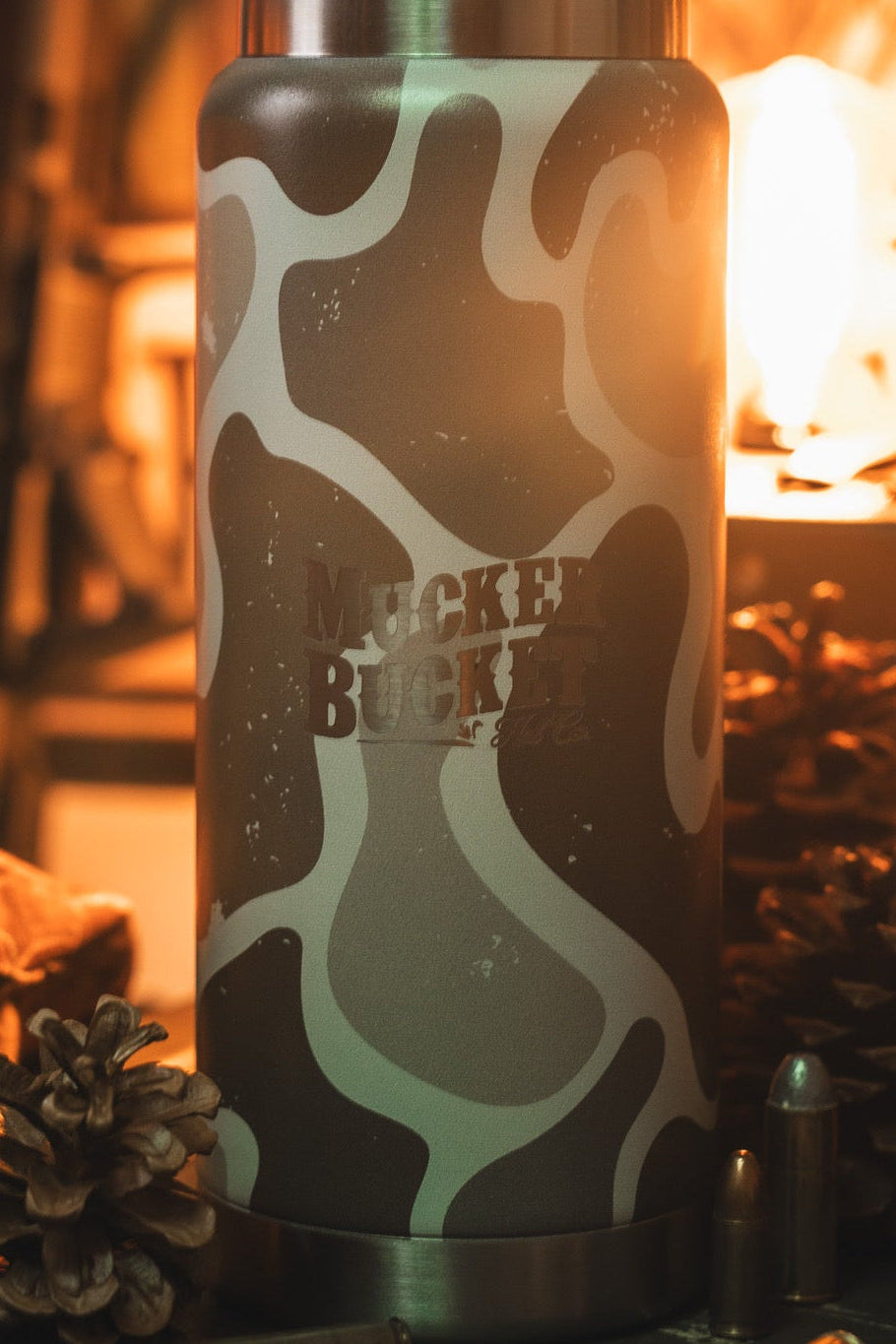 A camouflage-patterned insulated bottle labeled Muckey Bucket stands upright, surrounded by pine cones and bullets, with a warm light source in the background.