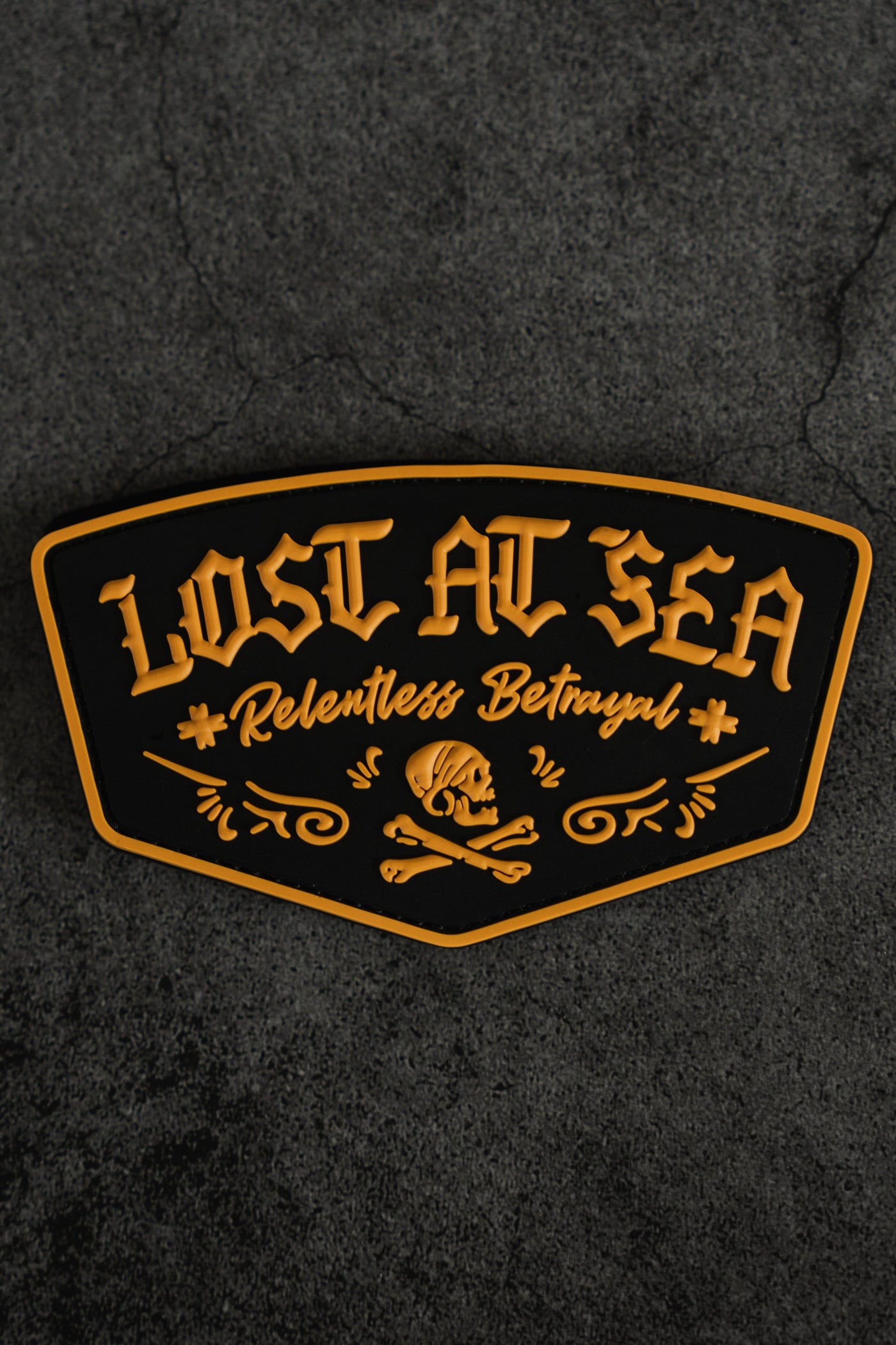A black and gold Lost At Sea PVC Patch by Relentless Betrayal, featuring a bold skull and crossbones design on a textured dark background—the perfect accessory for your Lost at Sea hoodie or survival hoodie.