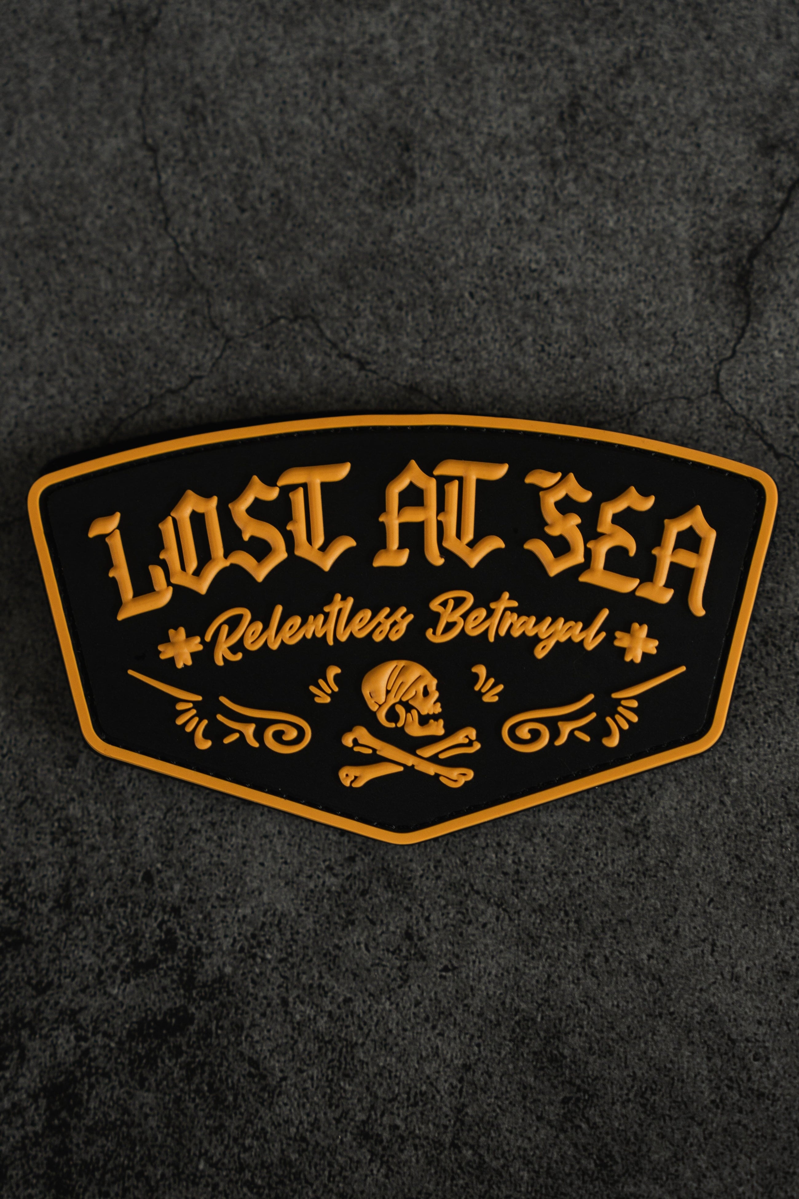 A black and gold Lost At Sea PVC Patch by Relentless Betrayal, featuring a bold skull and crossbones design on a textured dark background—the perfect accessory for your Lost at Sea hoodie or survival hoodie.