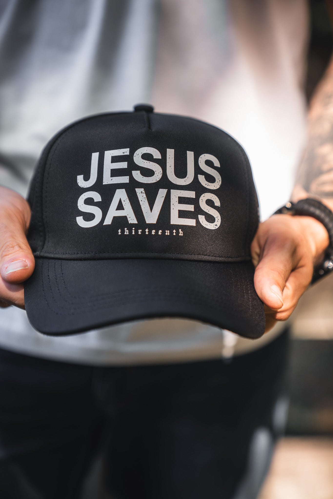 A person holds a black baseball cap with the words JESUS SAVES printed in large white letters and a smaller word thirteencnth below.