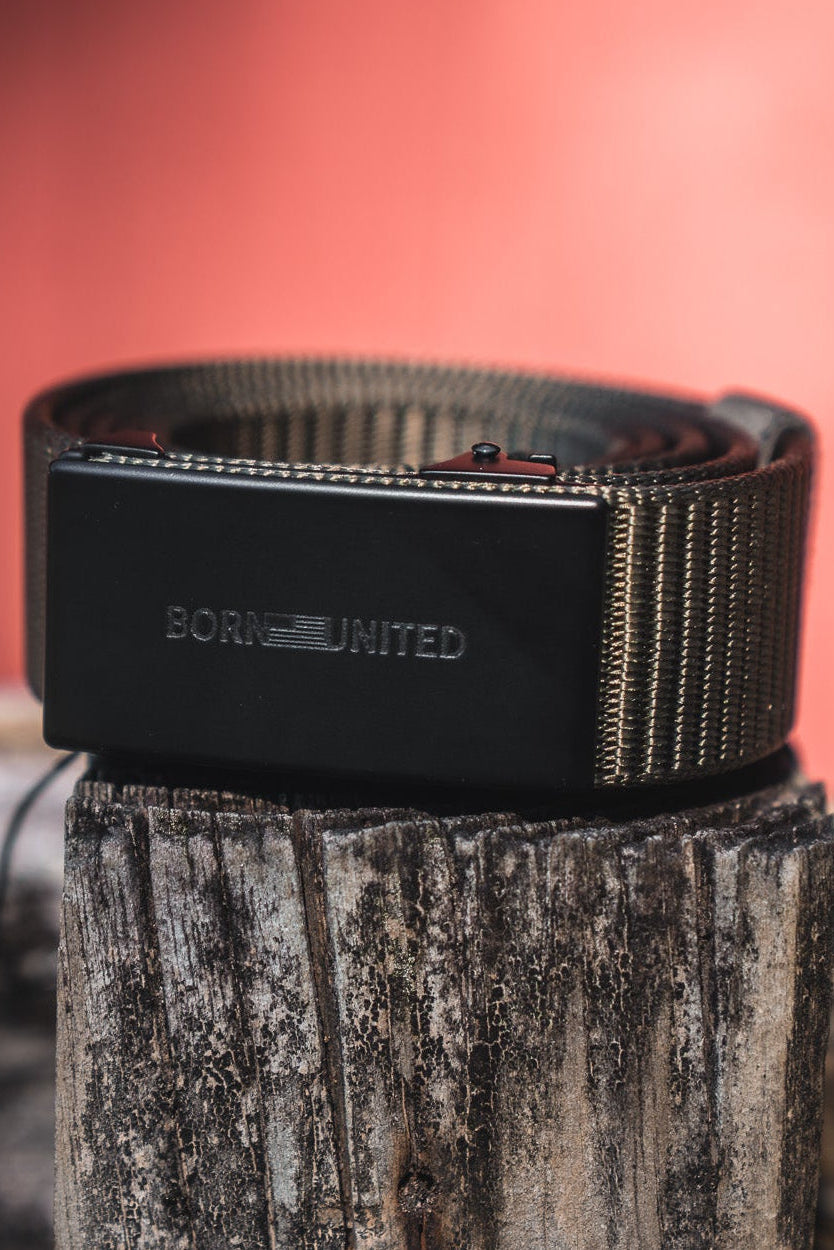 A close-up of a black belt with BORN UNITED embossed on the buckle, coiled and resting on a weathered wooden surface, with a blurred red background.