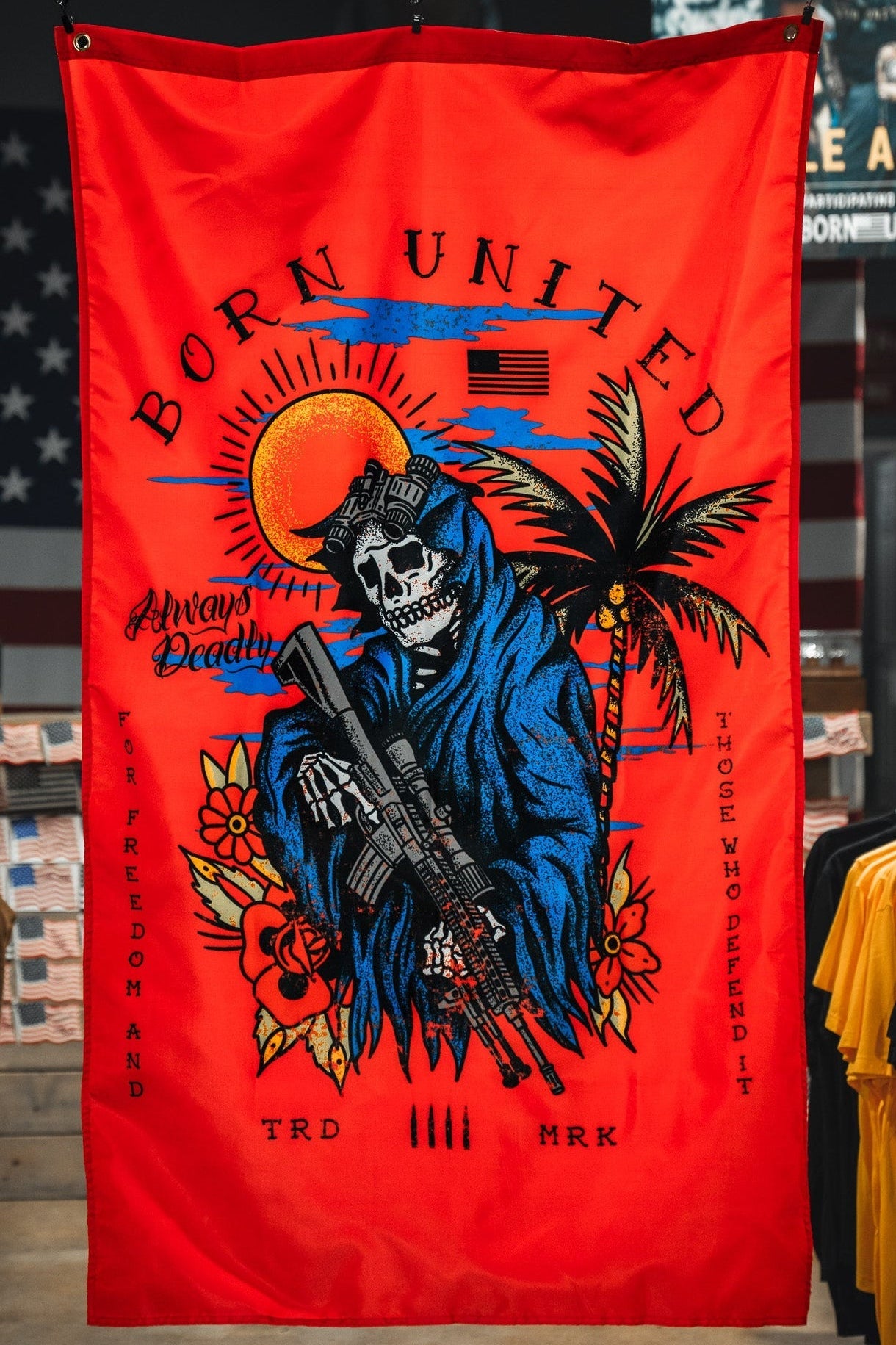 A red flag with a grim reaper skeleton holding a rifle, wearing tactical gear, set against a sunset, palm tree, and flowers. Text reads “Born United,” “Always Deadly,” and “For Freedom and Those Who Defend It.”.