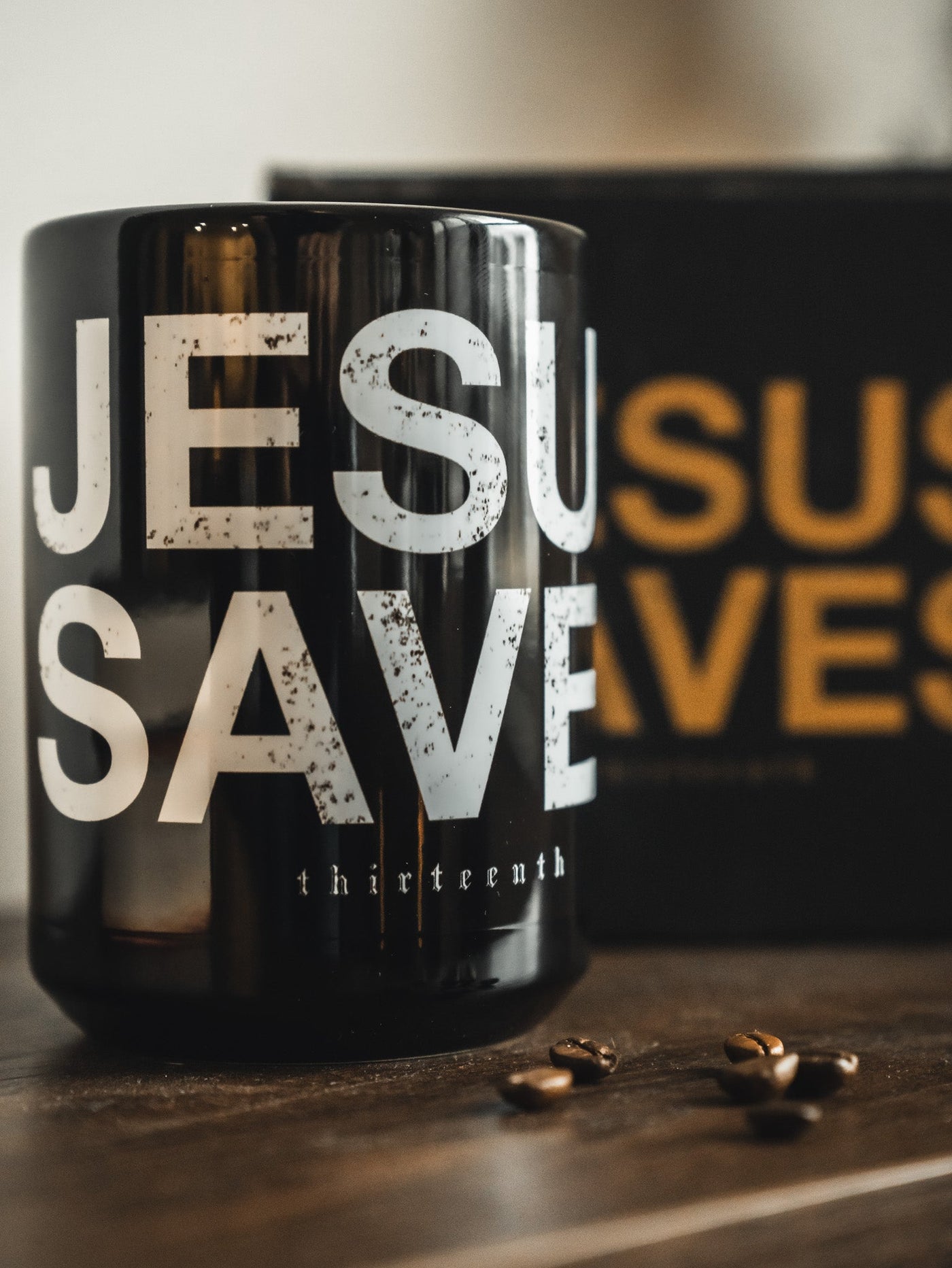 Black mug with large white text reading JESUS SAVES on a wooden surface, with coffee beans nearby; a blurred background shows yellow letters of the same phrase.