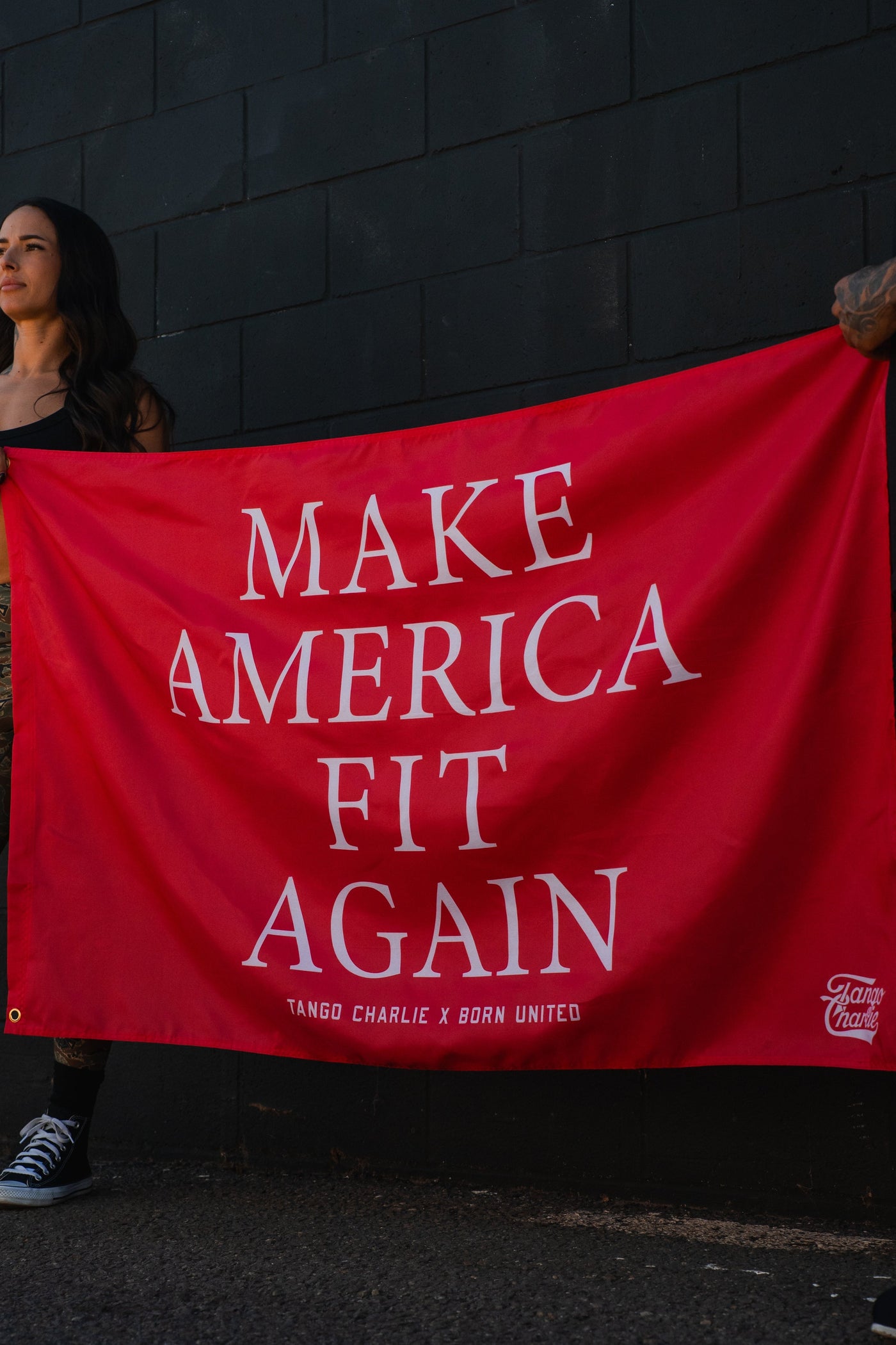 A red banner with white text reads MAKE AMERICA FIT AGAIN. People are partially visible holding the banner against a dark brick wall.