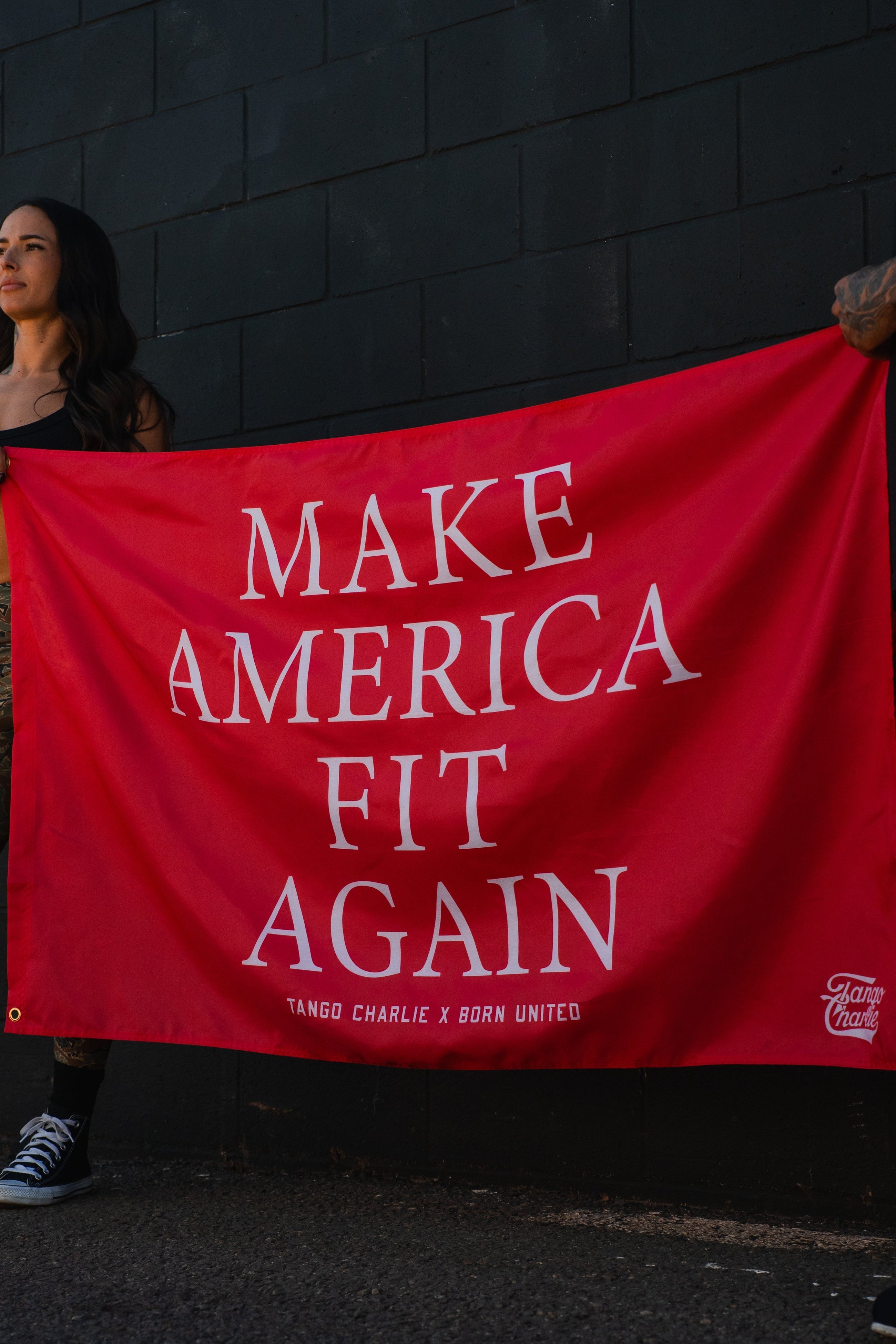 A red banner with white text reads MAKE AMERICA FIT AGAIN. People are partially visible holding the banner against a dark brick wall.