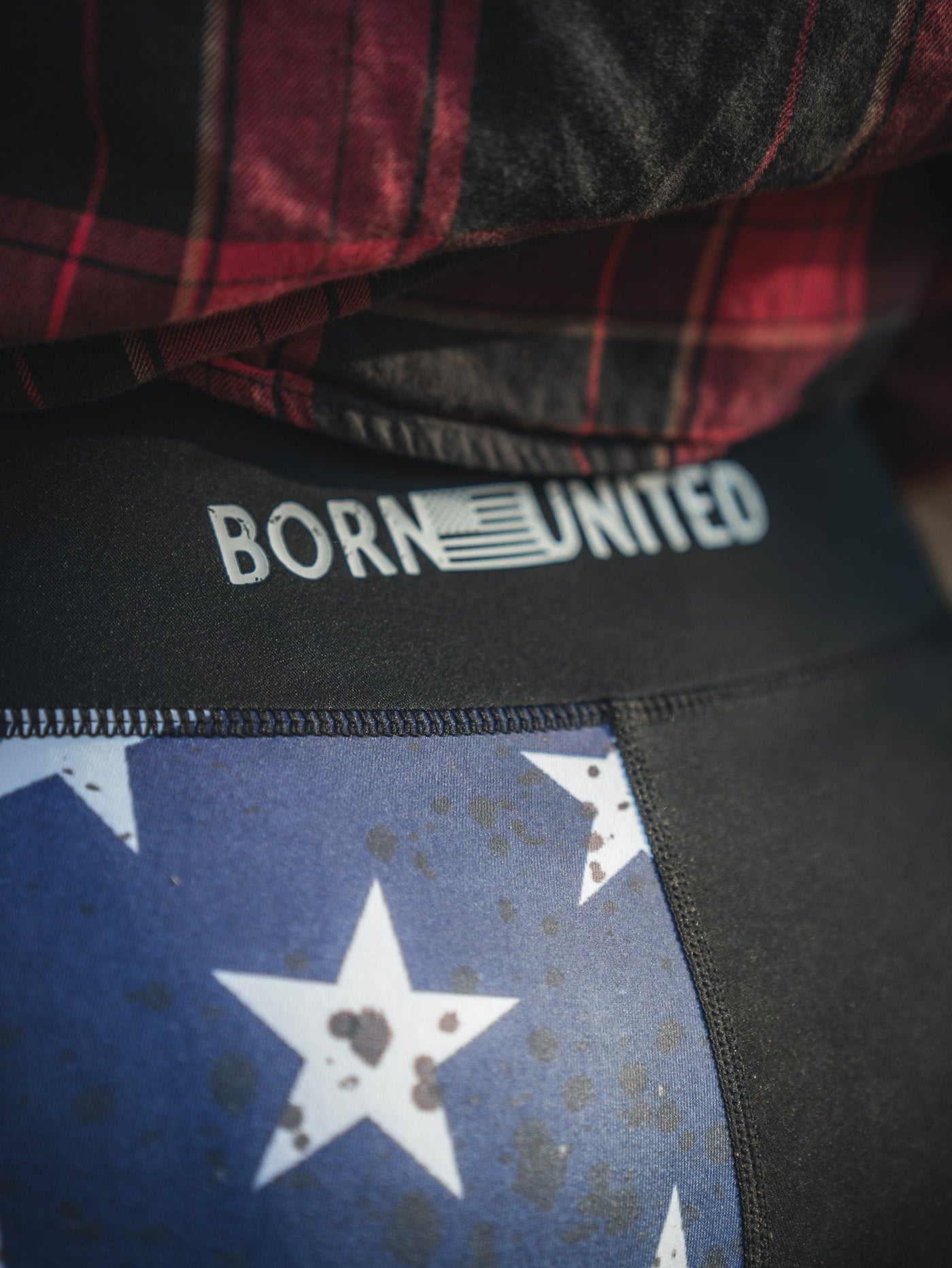 A person with tattooed hands wears black pants with BORN UNITED text and black-and-white sneakers, sitting on a brick ledge with hands resting in their lap.