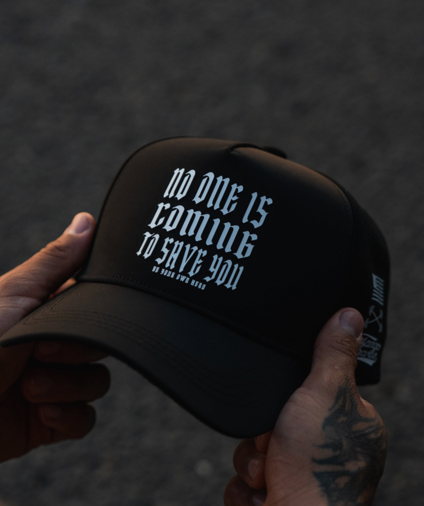 A tattooed person holds a black cap with white gothic text that reads, NO ONE IS COMING TO SAVE YOU against a blurred, dark background.