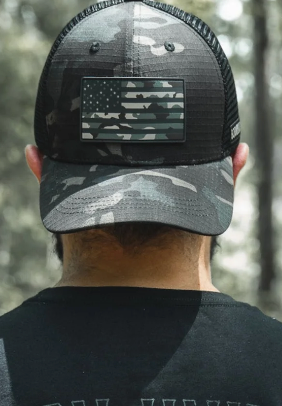 A person wearing a black and gray camouflage cap with a matching American flag patch, viewed from behind outdoors with blurred trees in the background.