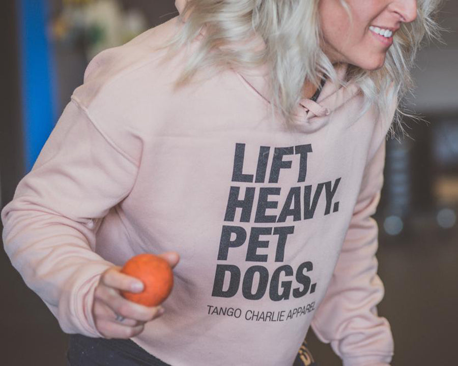 A person wearing a light pink hoodie with the text LIFT HEAVY. PET DOGS. holds an orange ball, smiling and looking to the side.