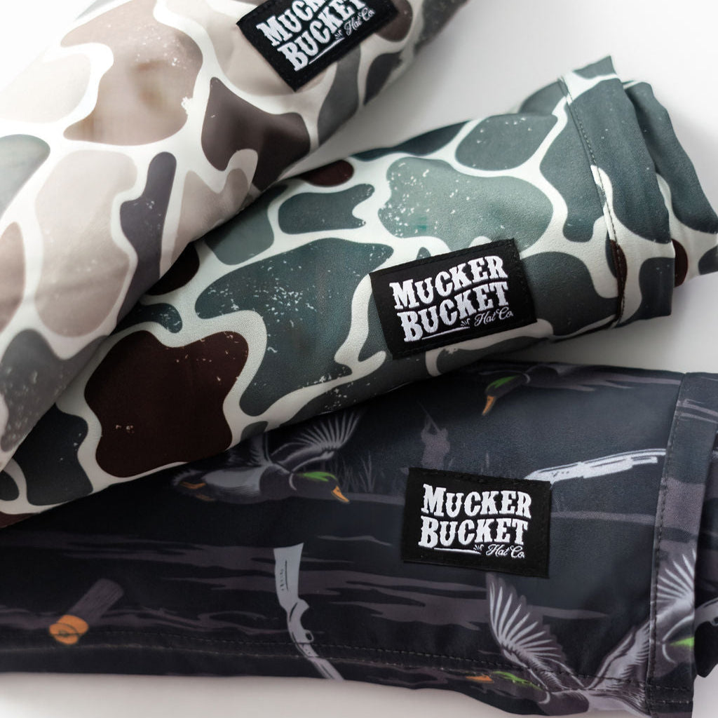 Three rolled-up fabric items with MUCKER BUCKET The Co. labels, featuring different camouflage and duck-themed patterns in shades of brown, green, and black.