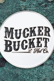 A round pin badge features Mucker Bucket in bold, decorative font on a textured teal and rust backdrop, capturing the vintage charm of an OG Duck Lucky Huntin Coin.