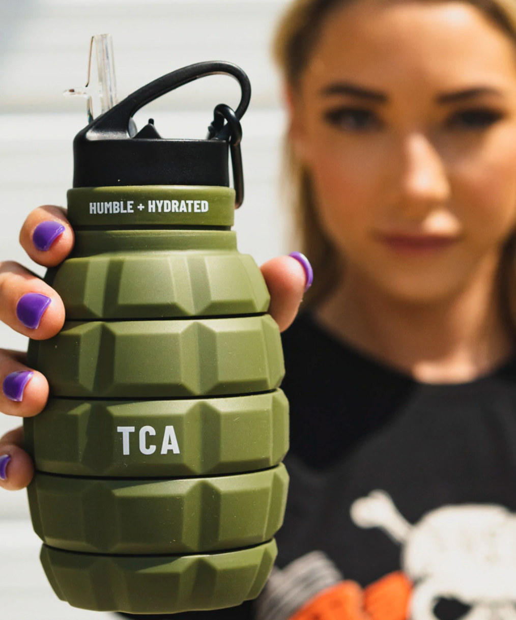 A woman with purple nail polish holds a green, grenade-shaped water bottle labeled HUMBLE + HYDRATED and TCA toward the camera, with her face blurred in the background.