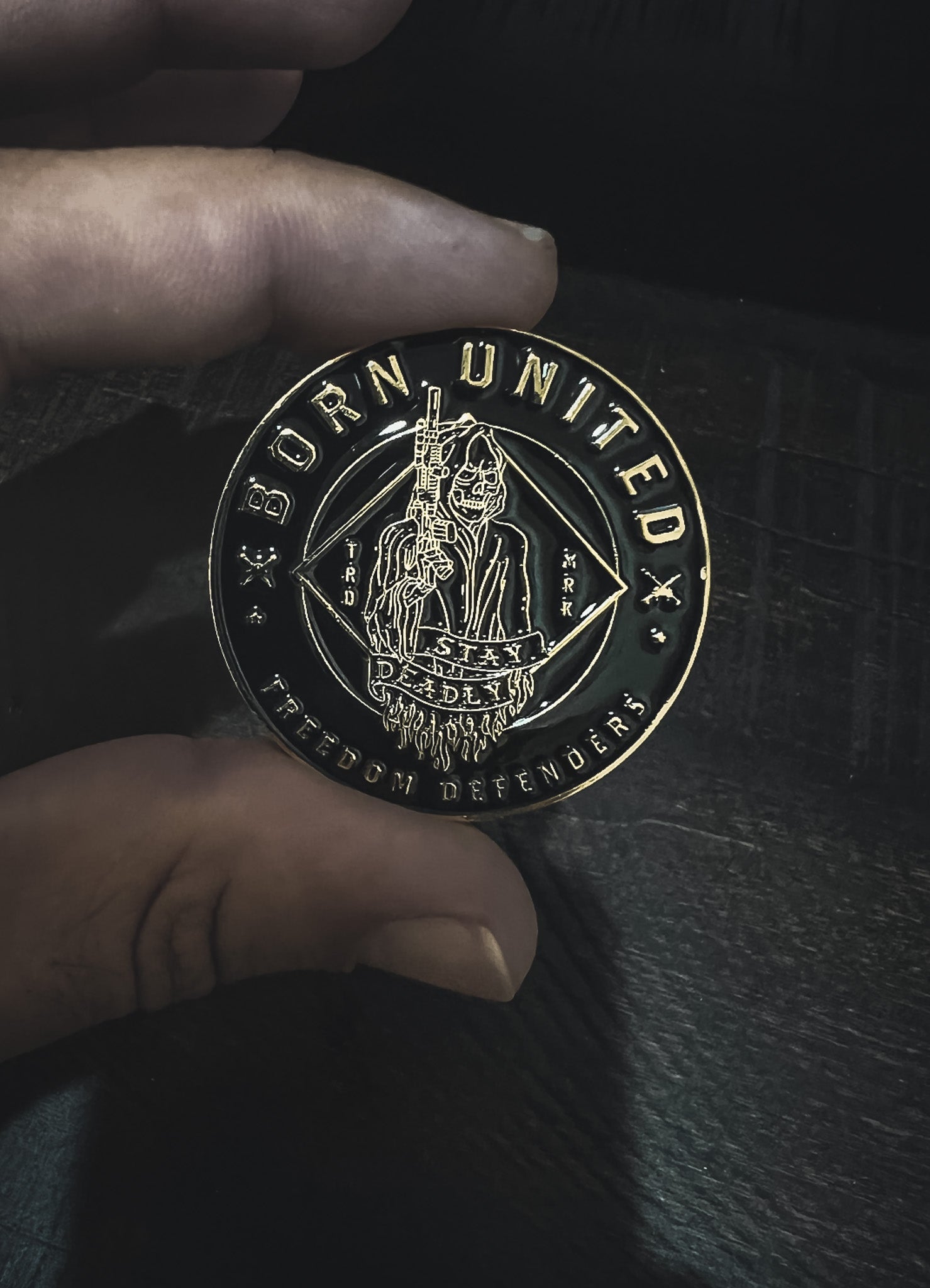 A hand holds a round metal coin with an engraved Grim Reaper holding a rifle, surrounded by text reading BORN UNITED and FREEDOM DEFENDERS against a dark background.