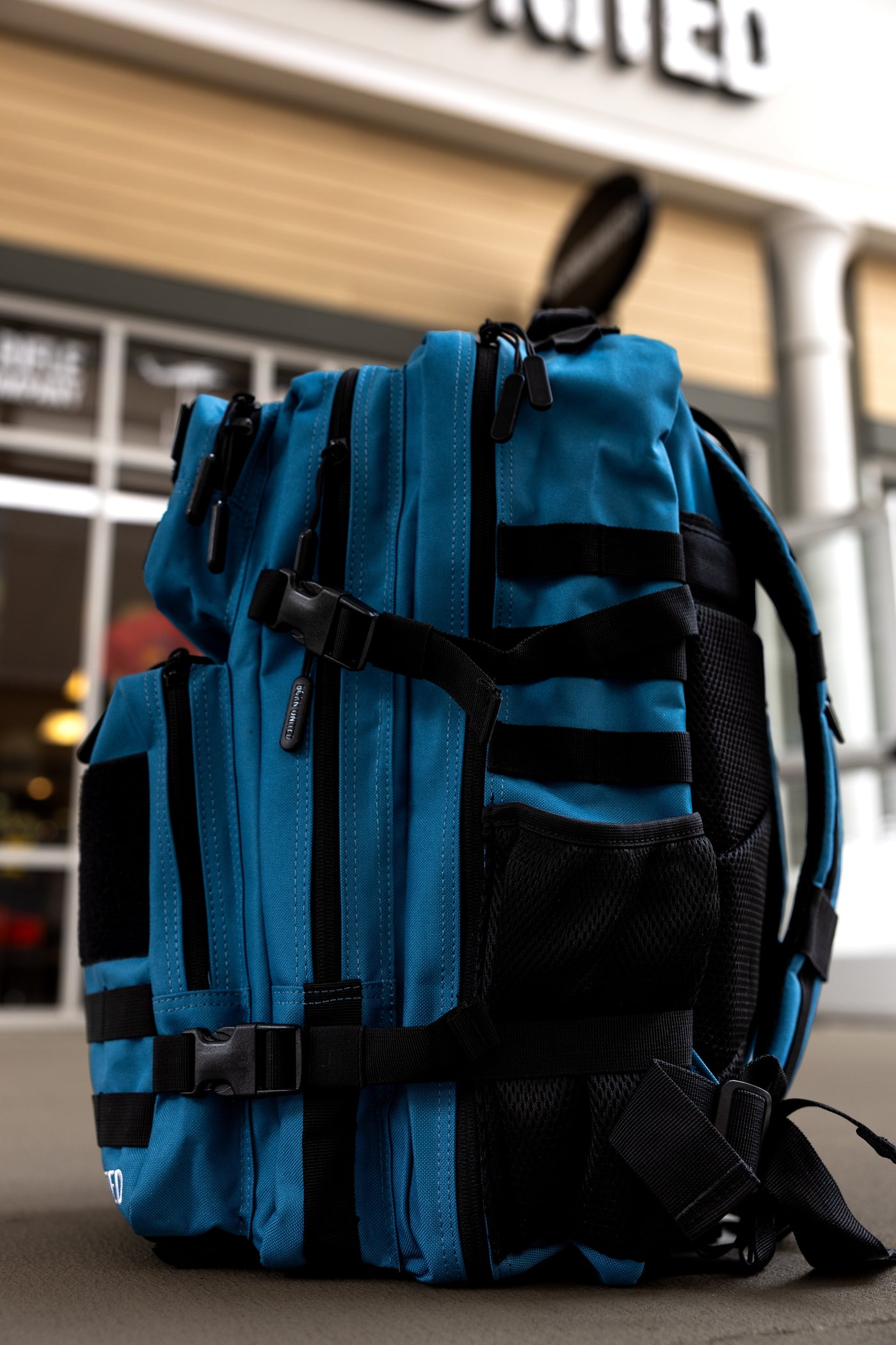 The Born United Private Label Peacock Blue Limited Ed. Tac-Pac - 45L, featuring multiple zippers and mesh pockets, is placed on the ground in front of a building with large windows and a sign above the entrance.