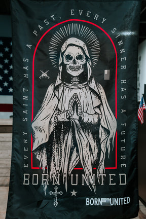 The Born United Private Label Saints and Sinners Flag is a black polyester flag featuring a skeletal saint in a robe with halo and praying hands, with the phrases EVERY SAINT HAS A PAST, EVERY SINNER HAS A FUTURE and BORN UNITED. An American flag appears in back.