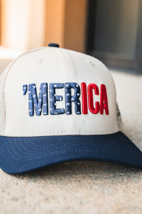 Introducing the Merica Trucker Snapback by Born United Private Label—a beige and navy snapback baseball cap featuring “MERICA” embroidered on the front, with “MER” in blue adorned with stars and “ICA” in bold red letters. This breathable, handmade hat offers a unique blend of comfort and style, making it perfect for fans of distinctive headwear.