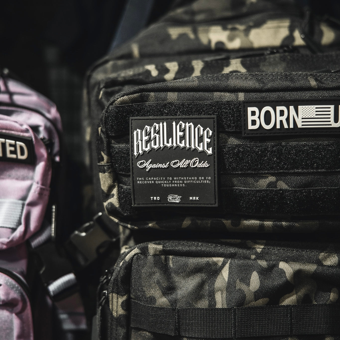 A close-up shows a camouflage backpack with the Tango Charlie Apparel "Resilience - Patch" and a 3D PVC American flag patch reading BORN. Part of a pink bag appears on the left.