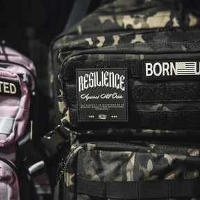 A close-up shows a camouflage backpack with the Tango Charlie Apparel "Resilience - Patch" and a 3D PVC American flag patch reading BORN. Part of a pink bag appears on the left.