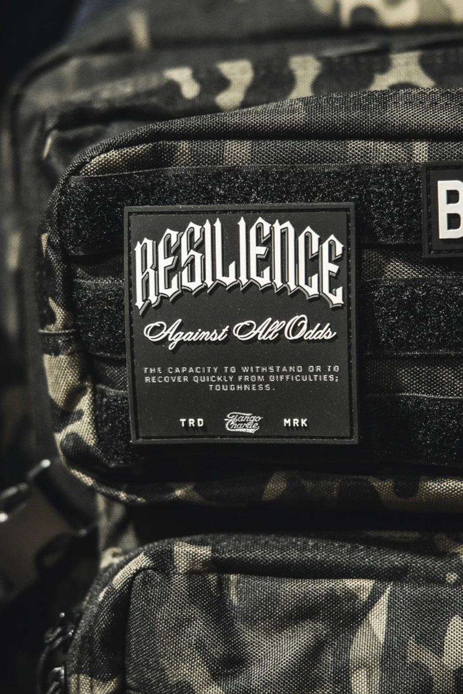 A close-up shows a camouflage backpack with the Tango Charlie Apparel "Resilience - Patch" and a 3D PVC American flag patch reading BORN. Part of a pink bag appears on the left.
