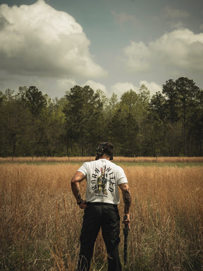 In a field of tall grass under a cloudy sky, a person stands facing away, holding a stick. They wear Born United Private Labels Battle Bunny white shirt featuring a unique design on the back, while tall trees line the horizon like guests at nature’s Easter dinner.