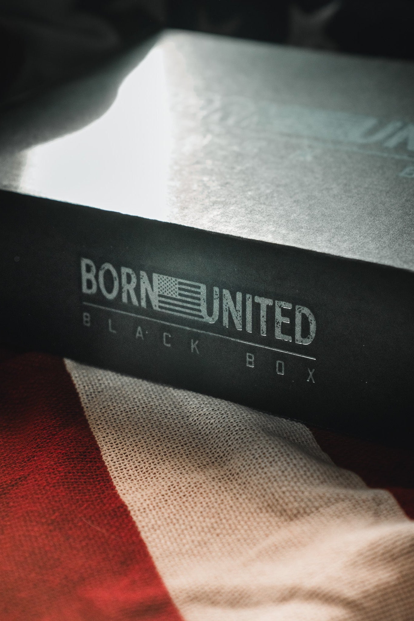 The Born United Private Label "Born United Black Box - Tactigator," featuring an American flag design, sits on a red and white striped cloth—inside are limited edition or mystery tees for collectors and fans.