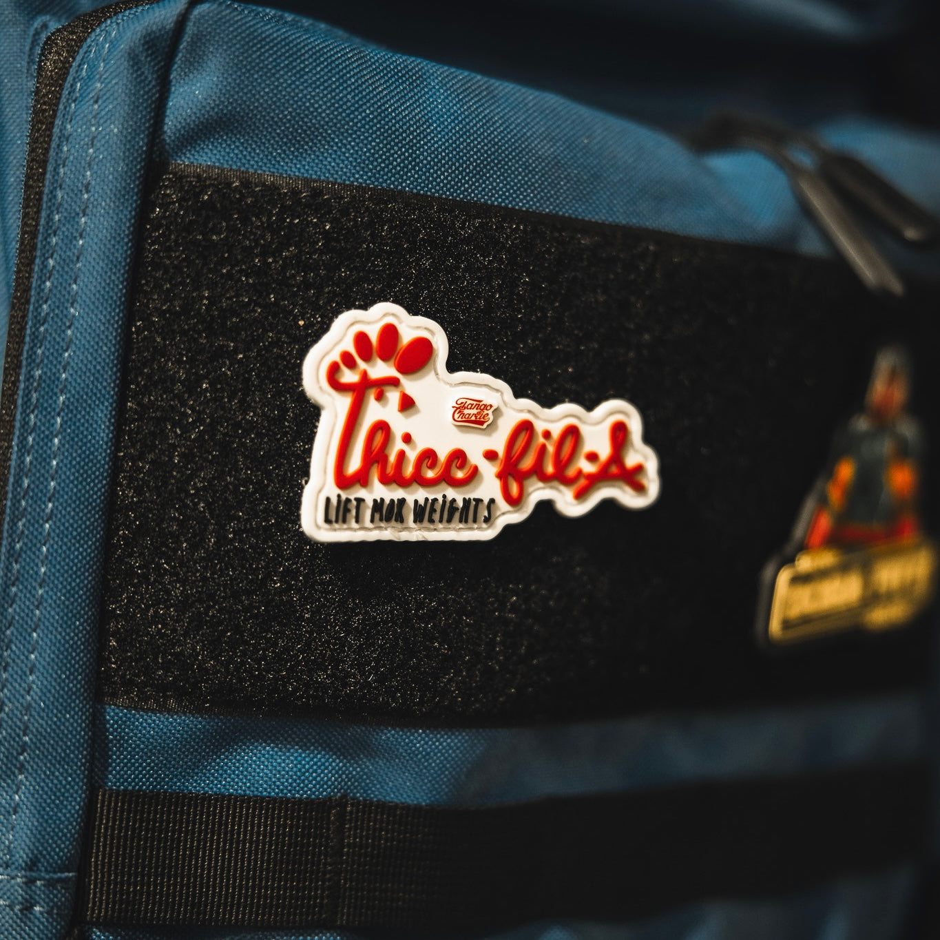A blue bag displays the Thicc-Fil-A Patch by Tango Charlie Apparel, featuring a 3-inch Velcro patch with "Thicc-fil-a Lift Mor Weights," a red paw print, and playful font attached to a black strip. Another patch appears partially on the right.