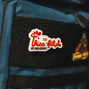 A blue bag displays the Thicc-Fil-A Patch by Tango Charlie Apparel, featuring a 3-inch Velcro patch with "Thicc-fil-a Lift Mor Weights," a red paw print, and playful font attached to a black strip. Another patch appears partially on the right.