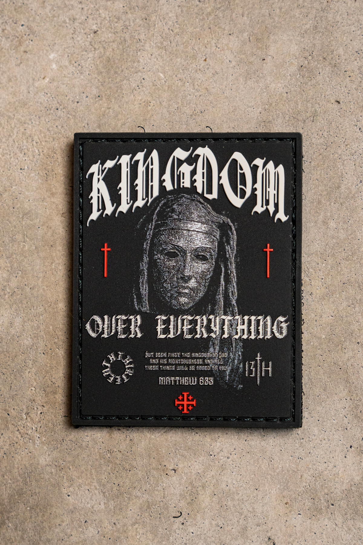 Kingdom Over Everything PVC patch, a 3-inch rectangular black design featuring a hooded figure, the text KINGDOM OVER EVERYTHING, small red crosses, and the Bible verse Matthew 6:33.